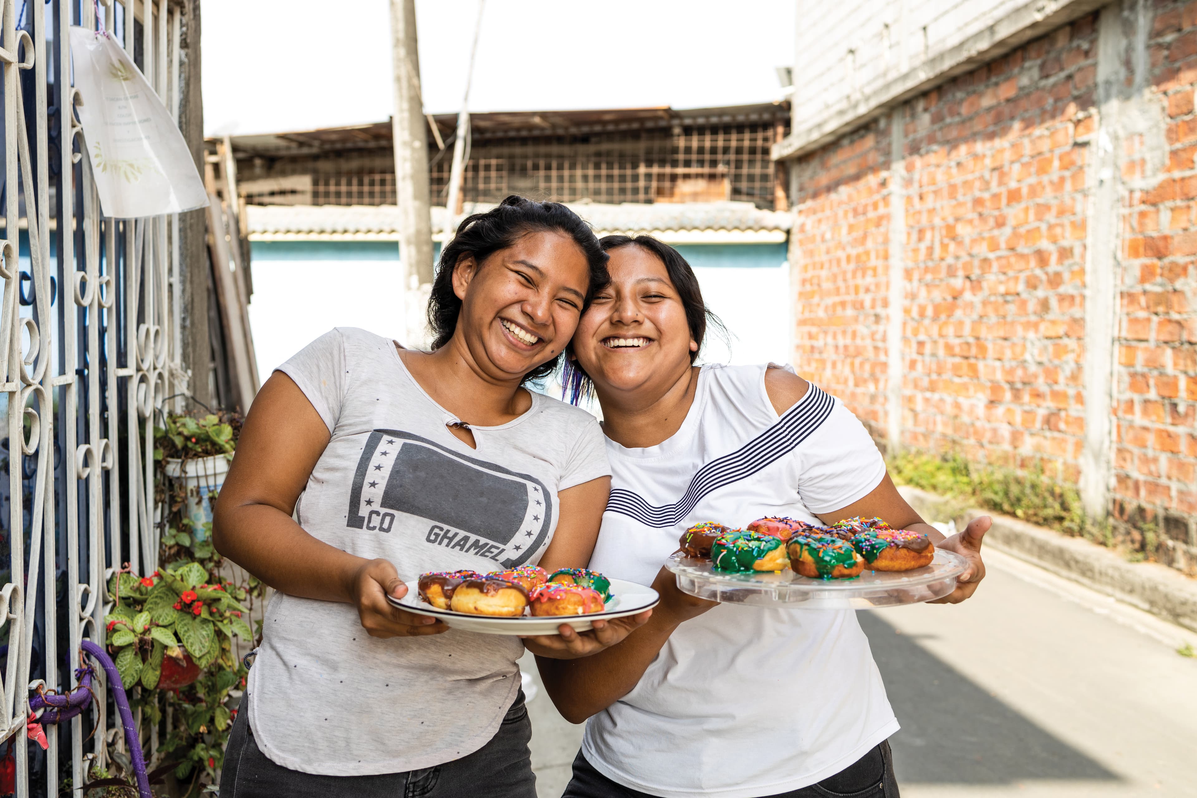Two young women stand outside holding plates of colorful donuts.
