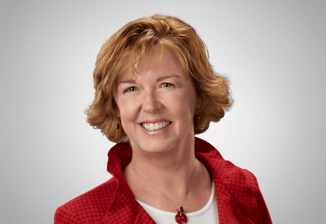 A woman in a white top, red blazer and red necklace is smiling, with a grey background behind her.