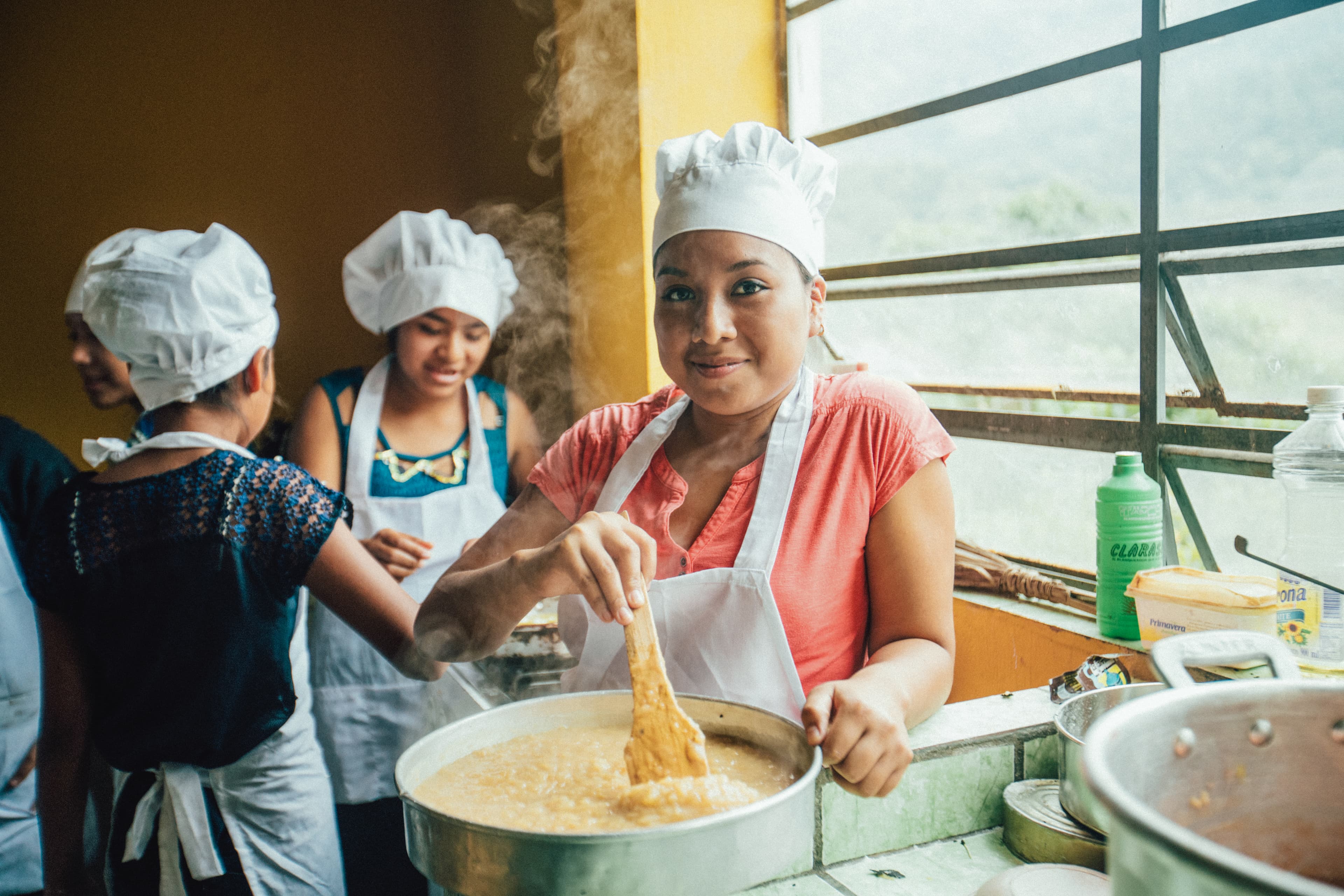 A teen wearing a white apron smiles while using a spoon and a bowl.