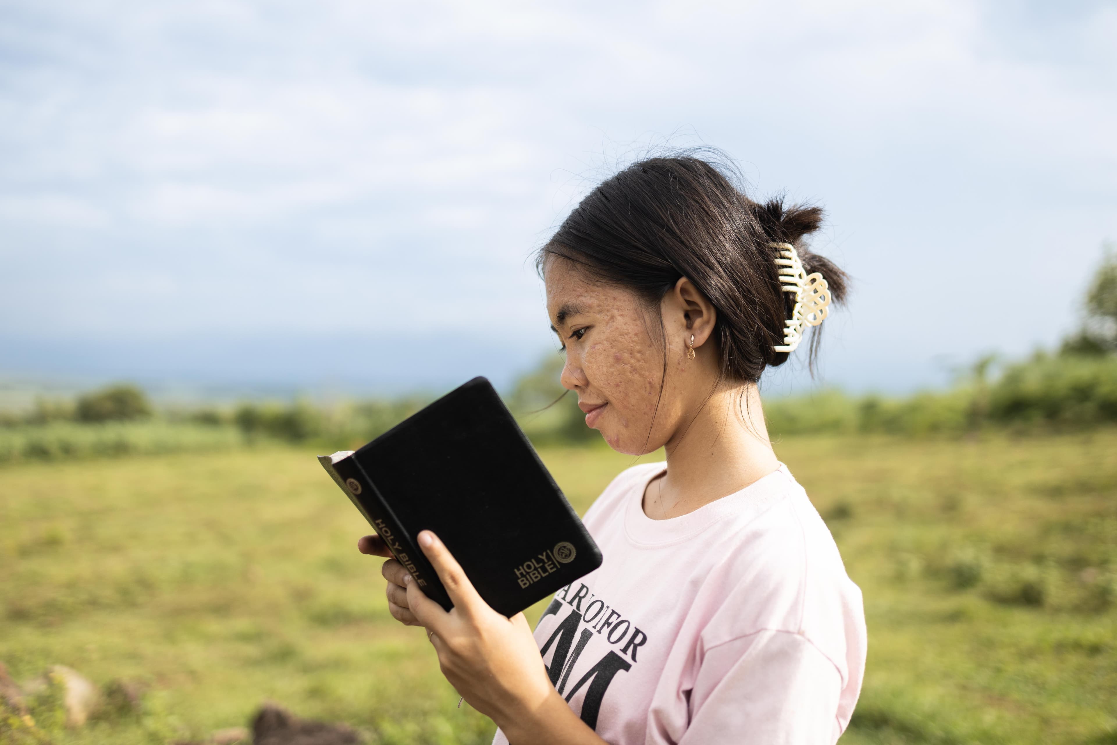 A teen girl stands in a field and reads a Bible.