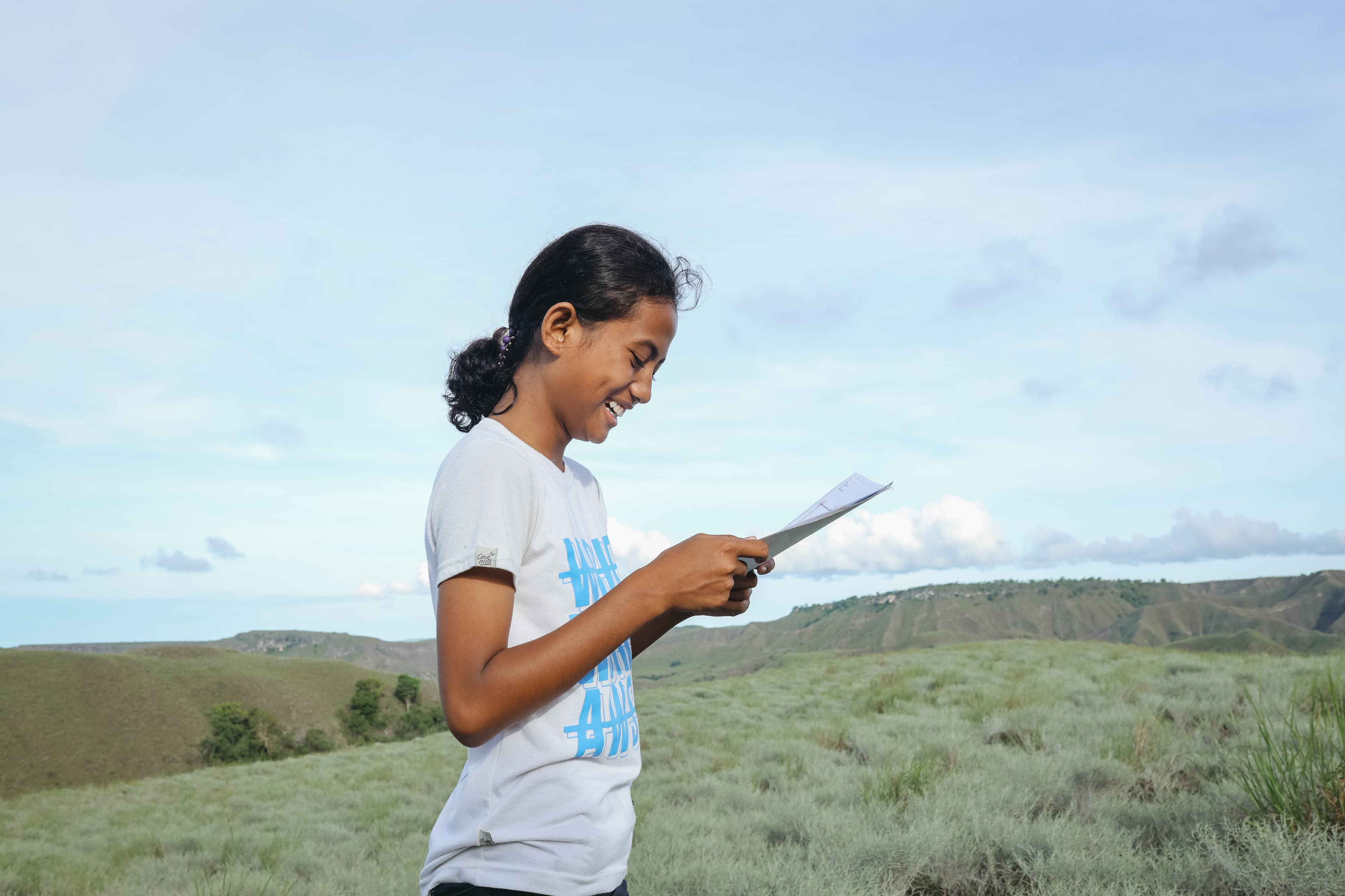 Girl wearing white T-shirt stands in field, smiles while reading a letter.