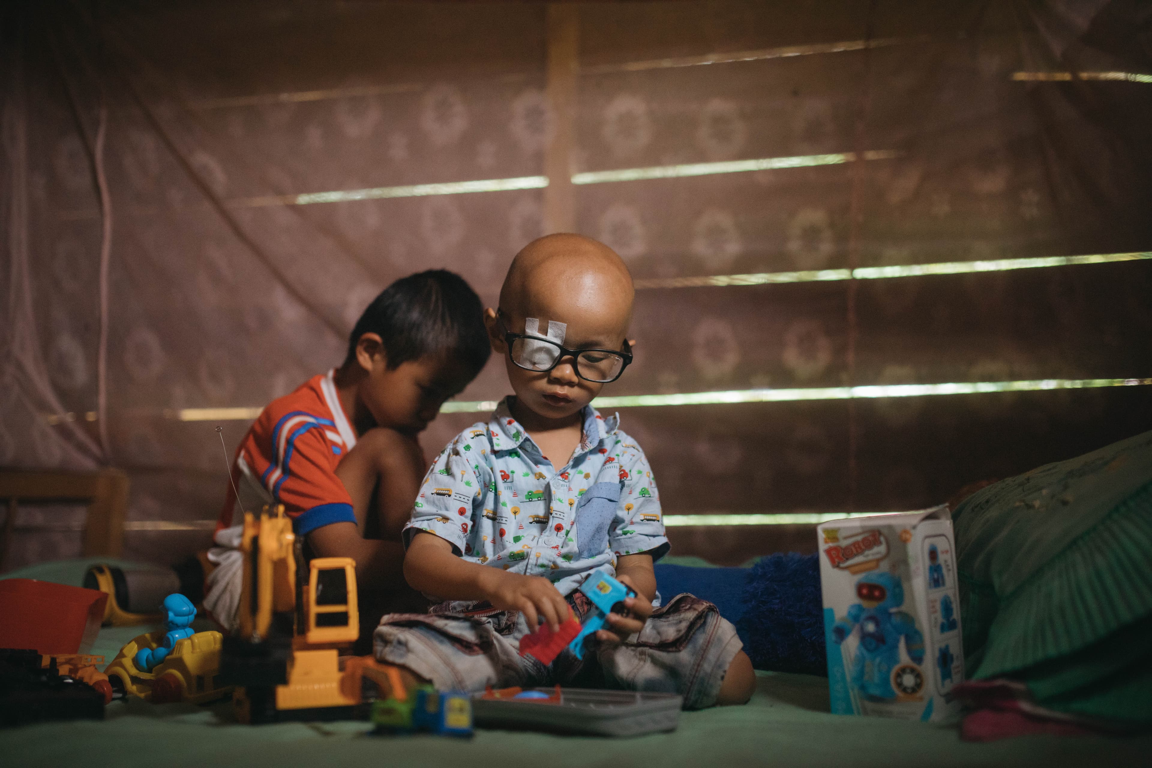 Young boy wearing eye patch and glasses sits, plays with toys alongside another child.