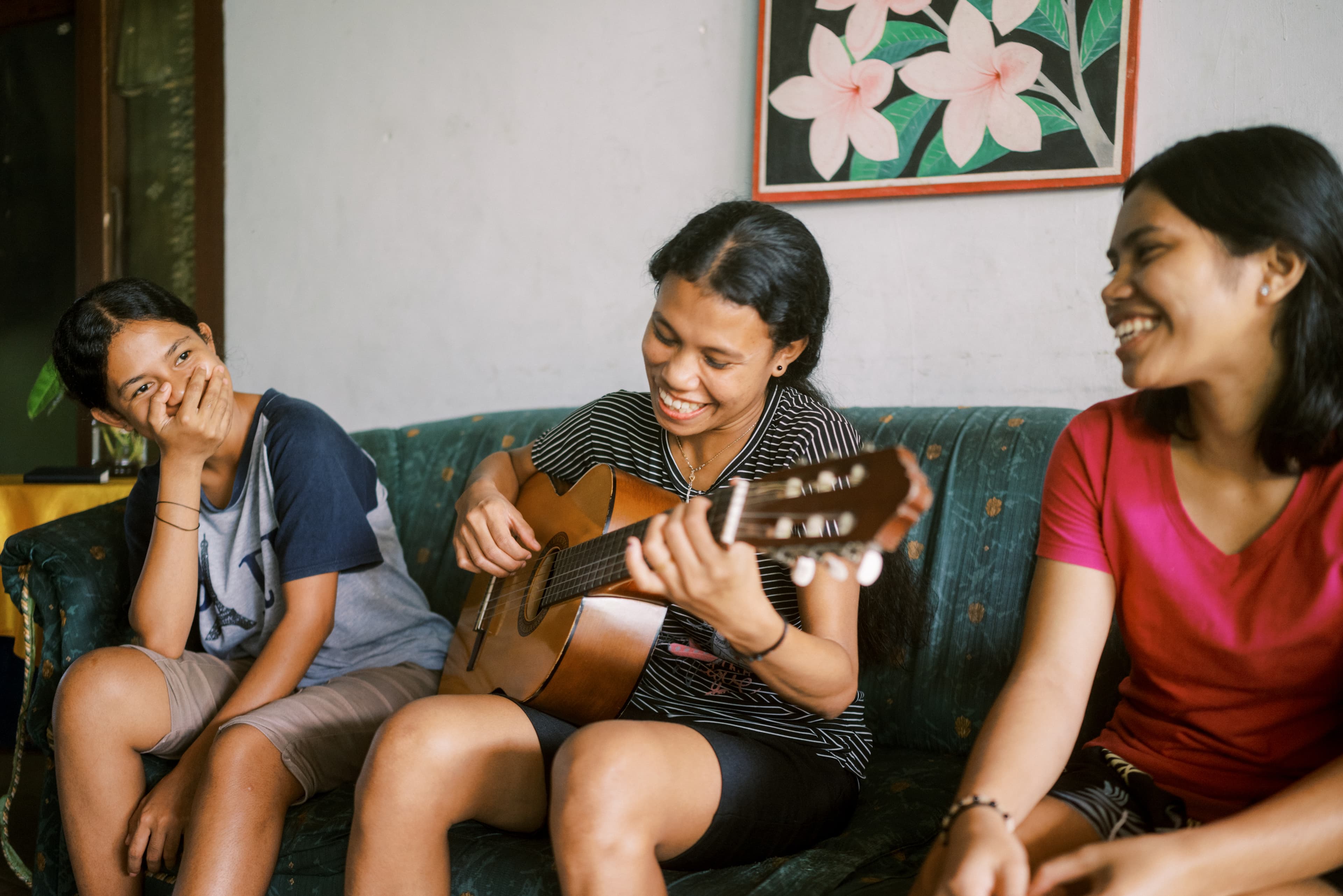 Three teen girls sit on a couch while one plays the guitar.