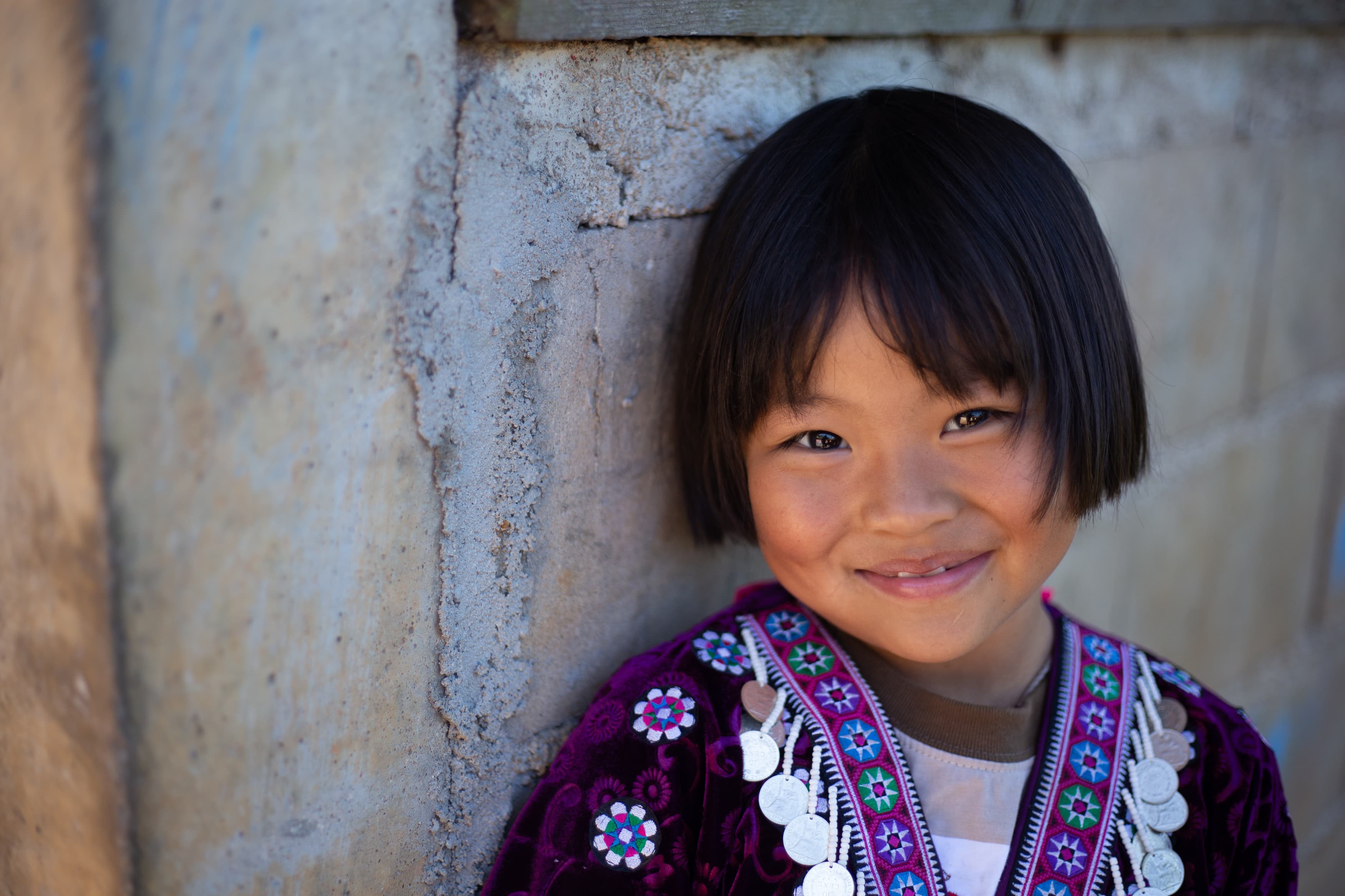 A little girl with dark hair leans against a wall and smiles brightly at the camera.