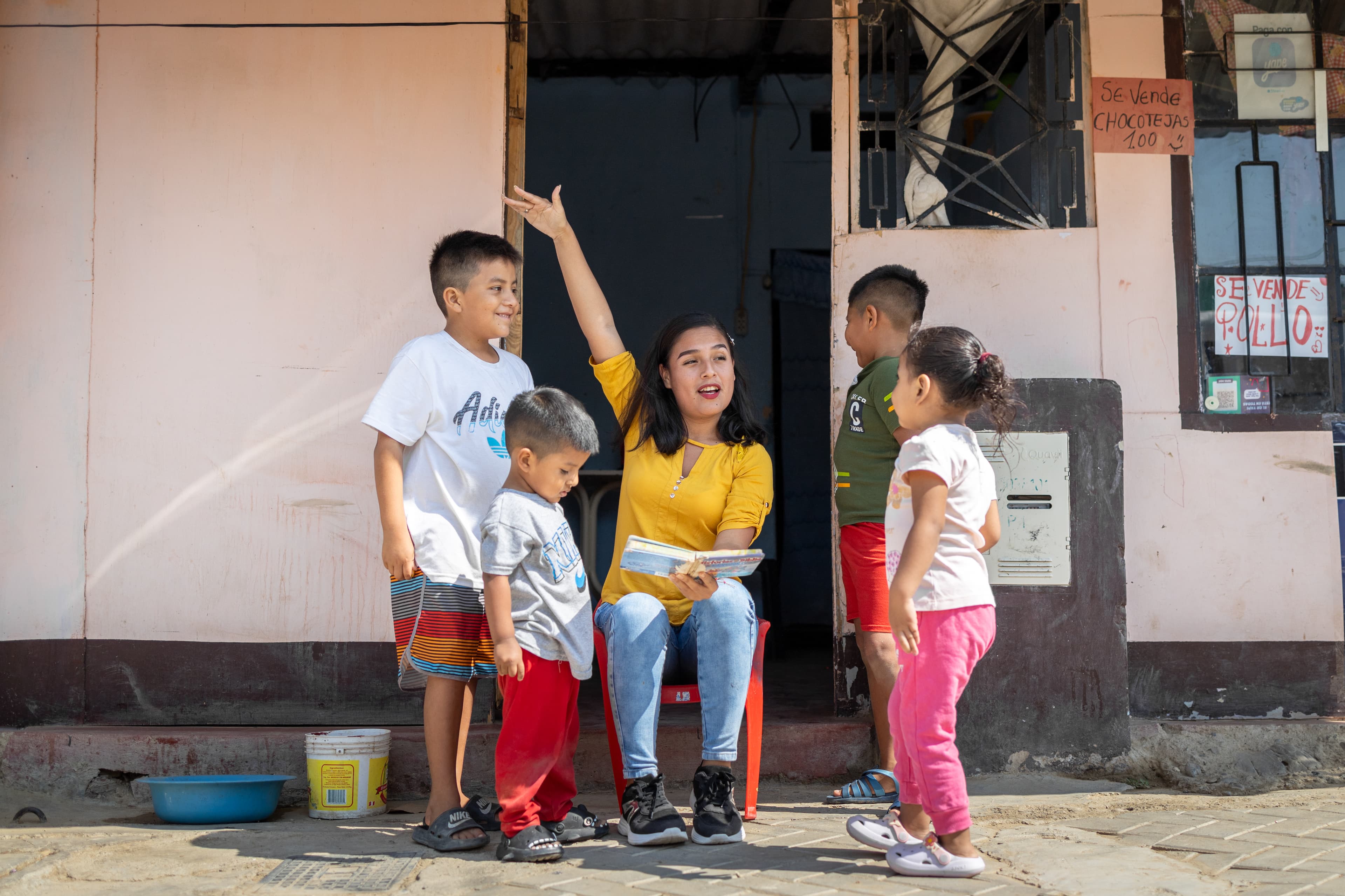 A young woman wearing a bright yellow shirt raises her arm while sharing a Bible story with the children around her.