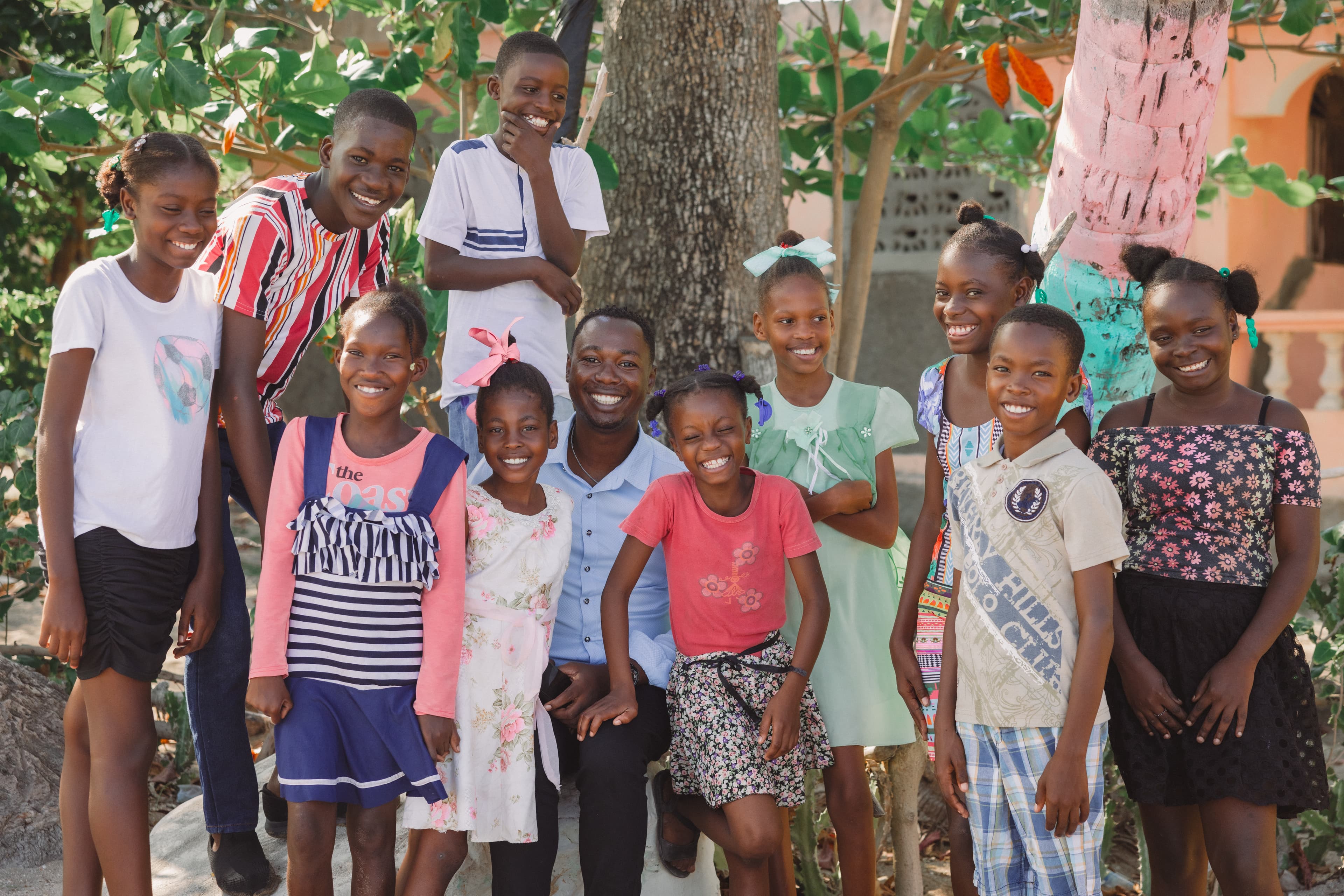 A young man sits in the middle of a group of children while smiling for the camera.