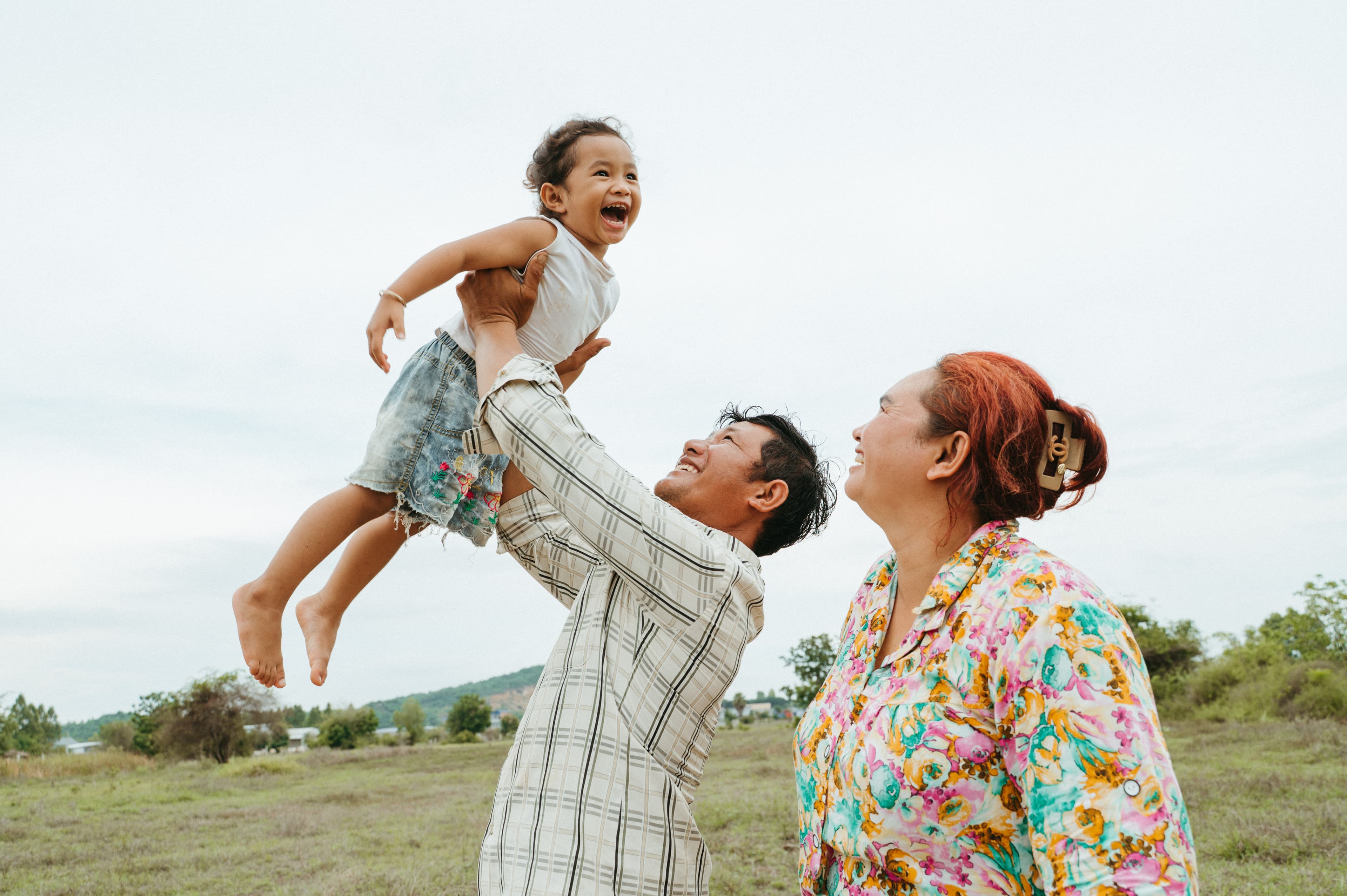 A father is playfully holding up his smiling daughter in the air as his wife stands next to him.