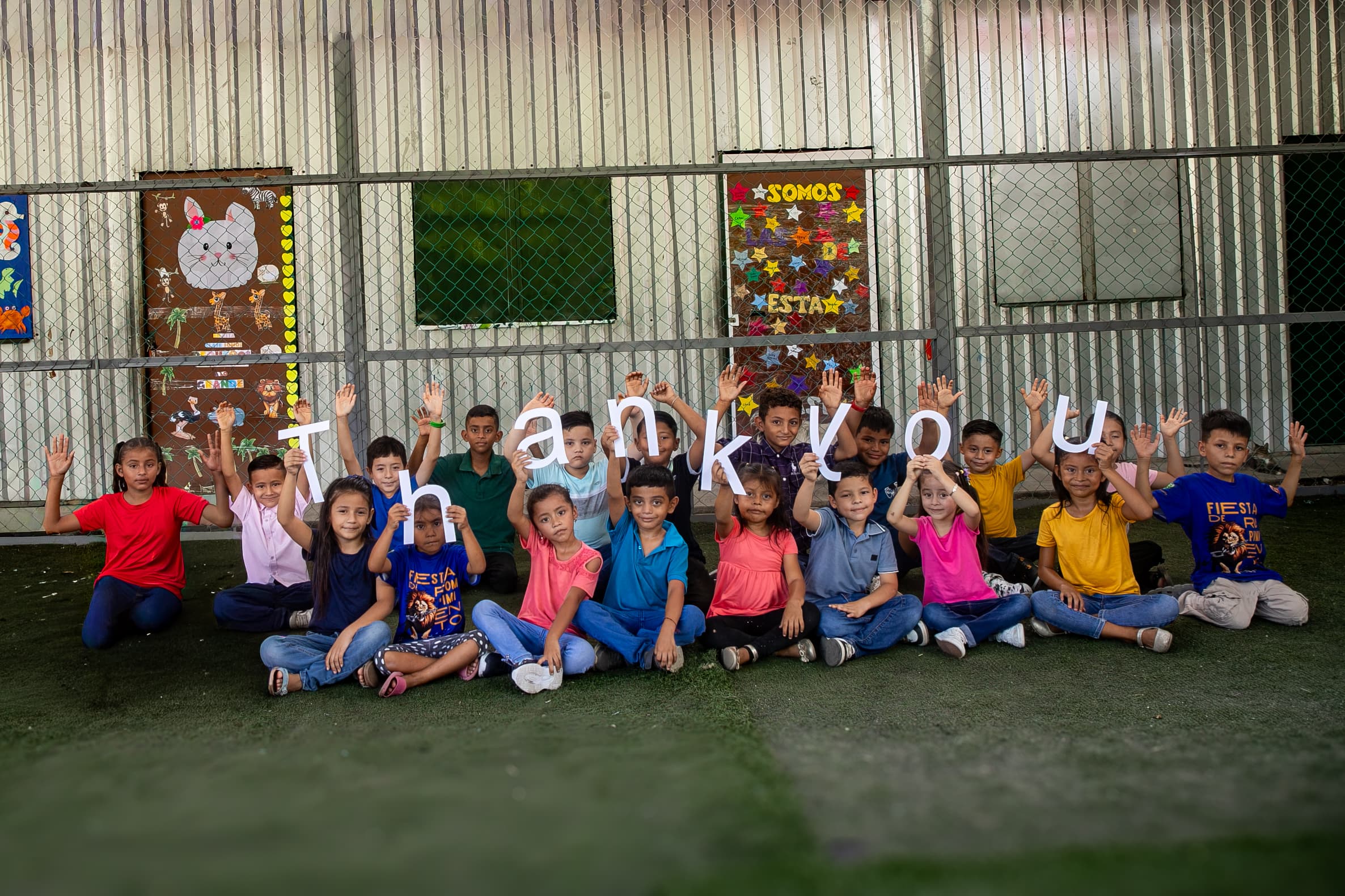 A group of children sit outside and hold up letters that spell out "Thank You".