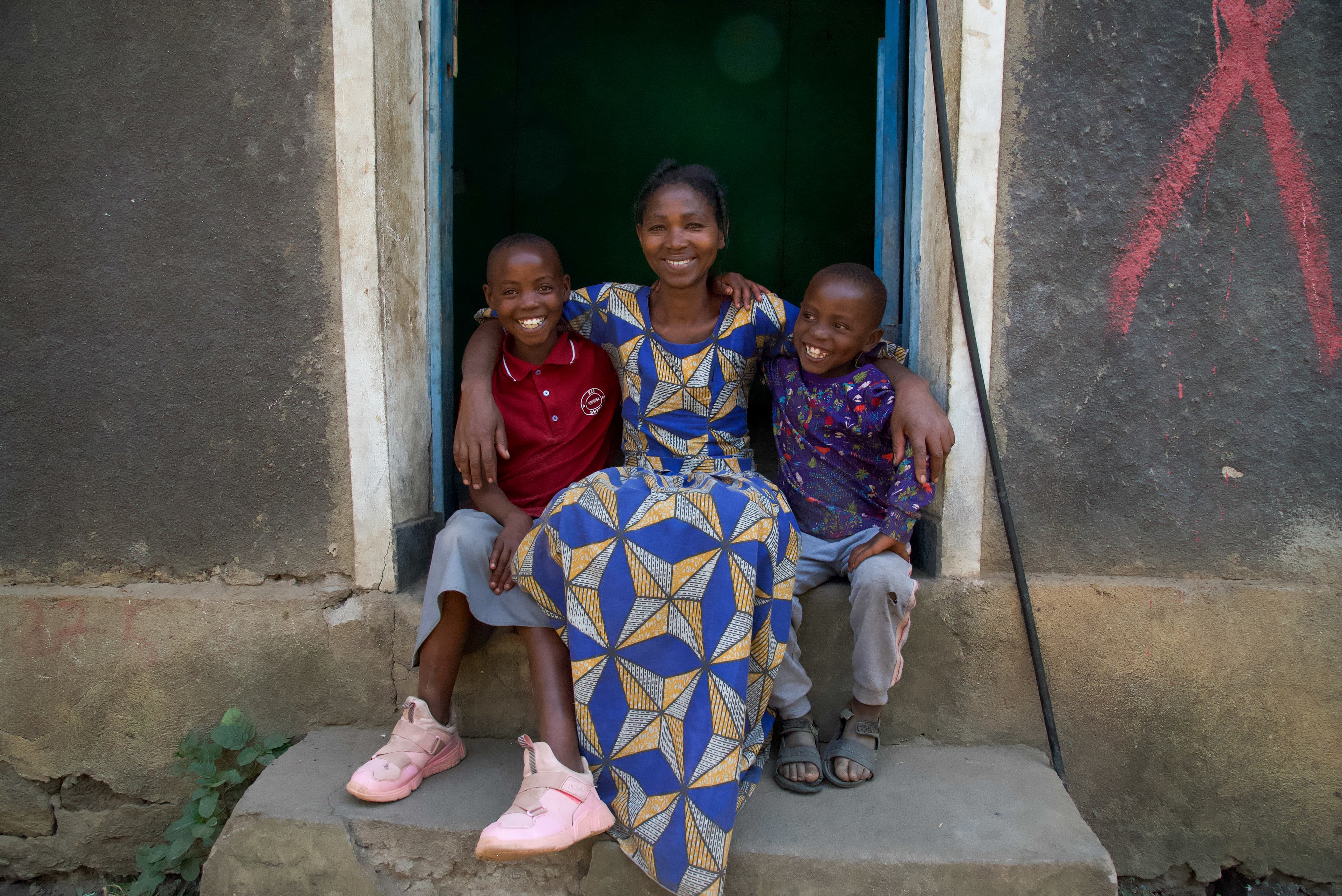 An African woman wearing a patterned dress sits in a doorway next to a young African girl and boy as they smile.