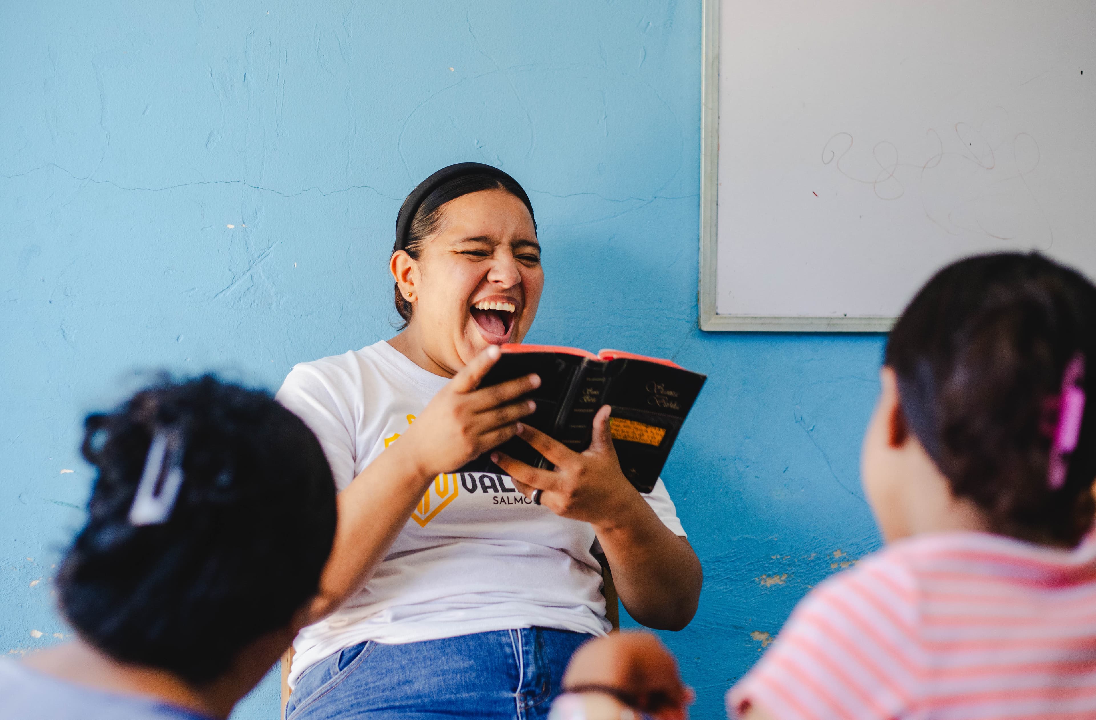A woman in a white t-shirt and jeans sits in a chair in a blue classroom, reading her Bible and laughing, with a whiteboard in the background.