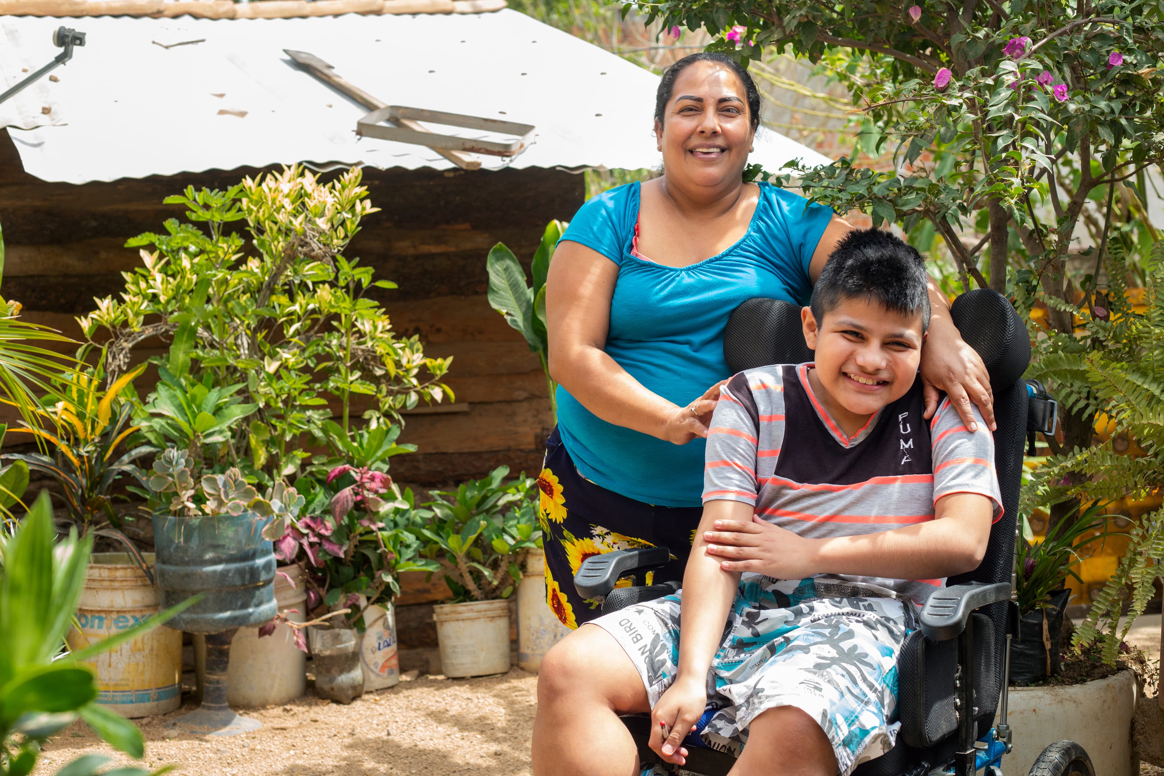 A young boy is sitting in a wheelchair smiling and his mother has her arms wrapped around him.