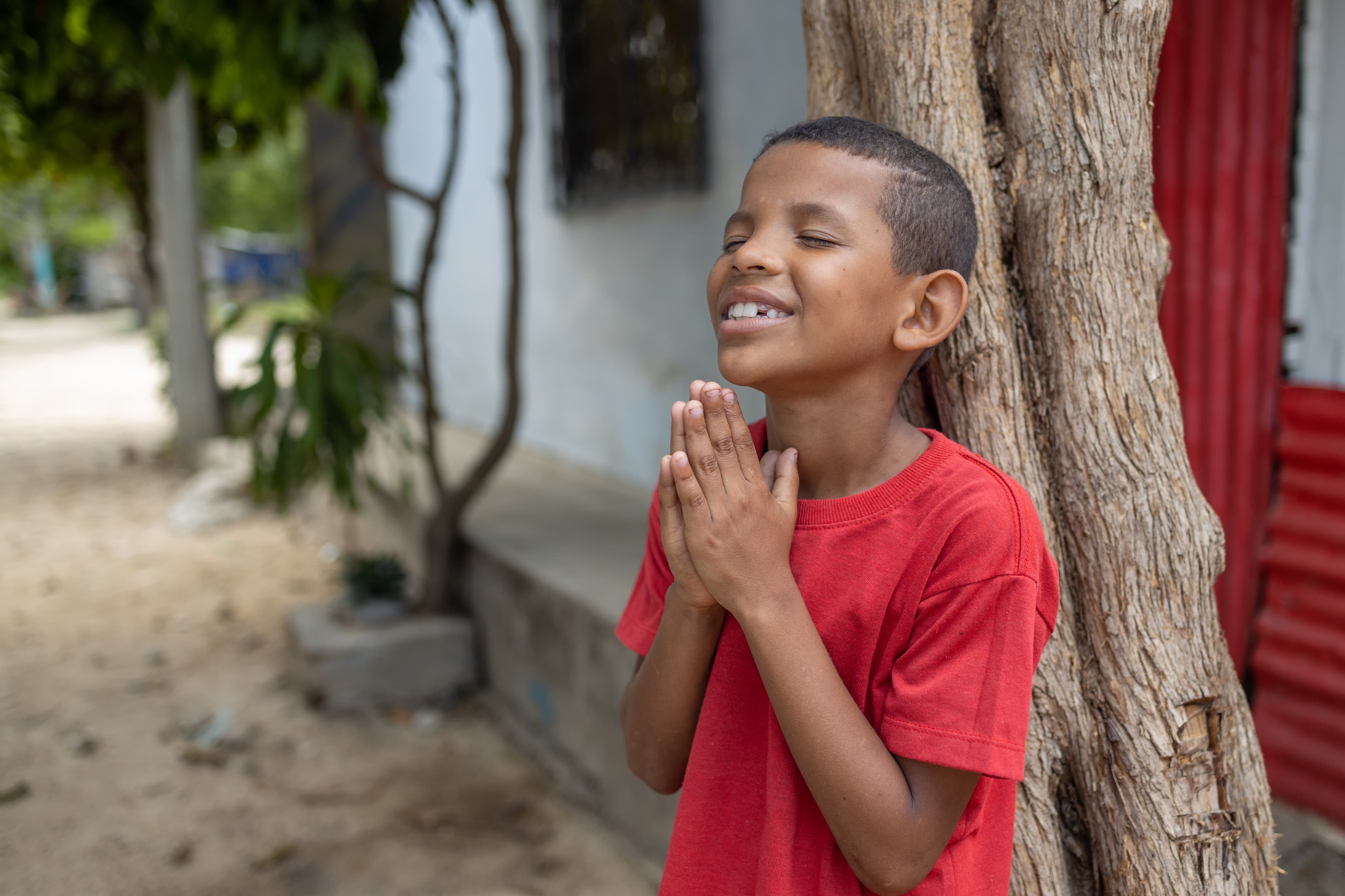 A Colombian boy wearing a red shirt leans against a tree with his hands folded in prayer.
