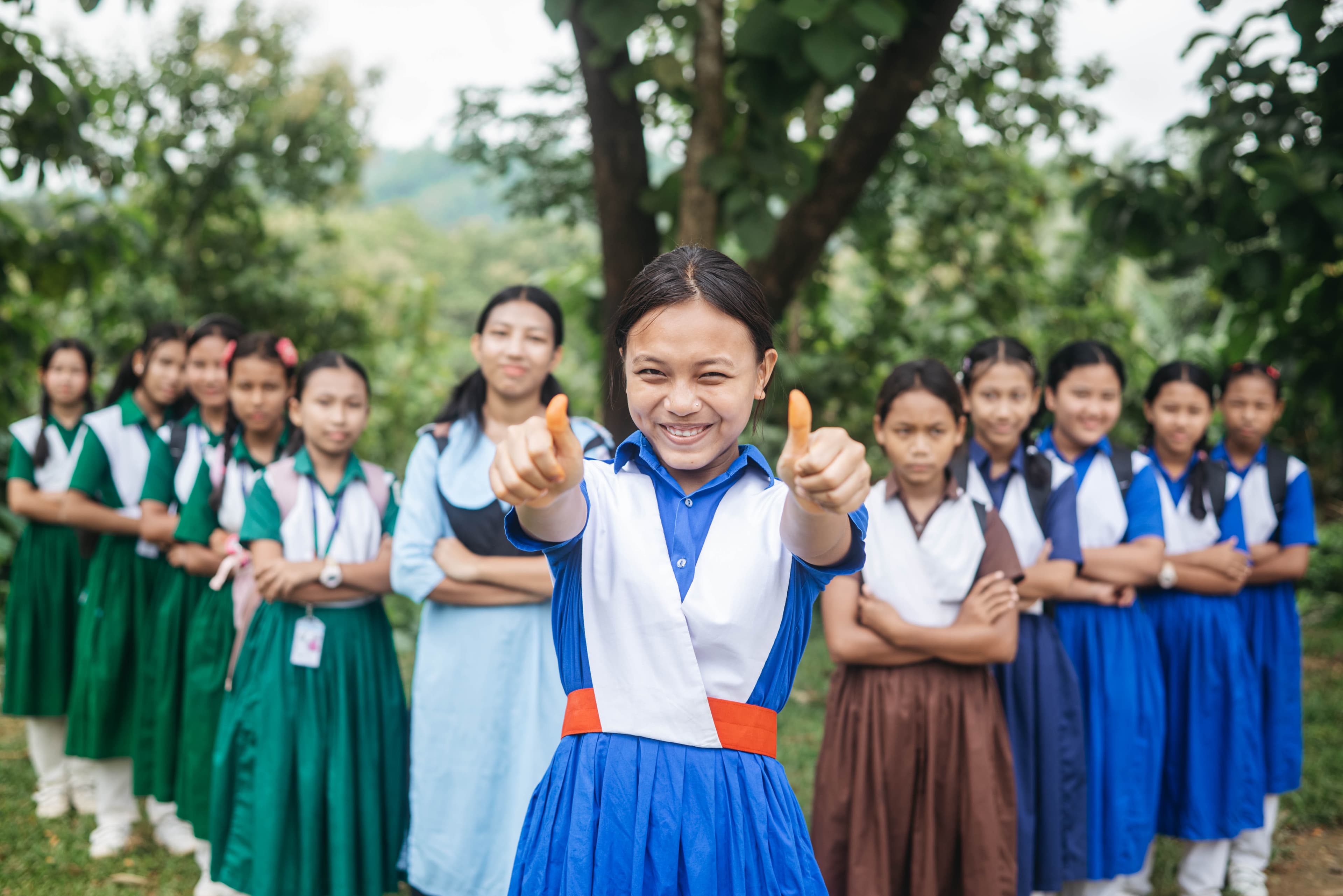 A teenage girl stands and smiles with her arms raised and her thumbs up as eleven other schoolgirls stand behind her in matching school uniforms, some of them different colors, and behind them are trees. .