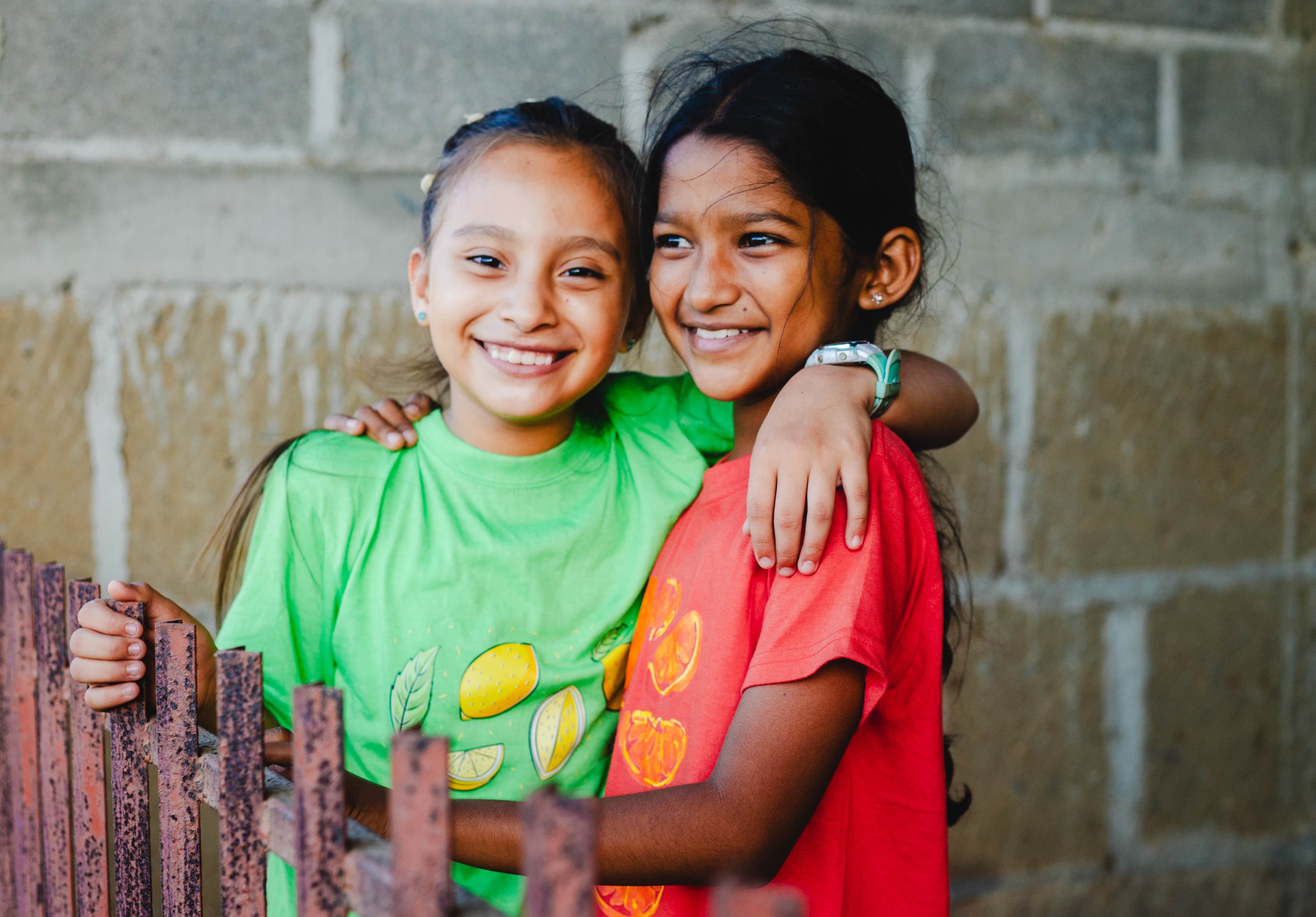 Two girls stand at a metal fence, embracing and smiling, with a cement brick wall in the background.