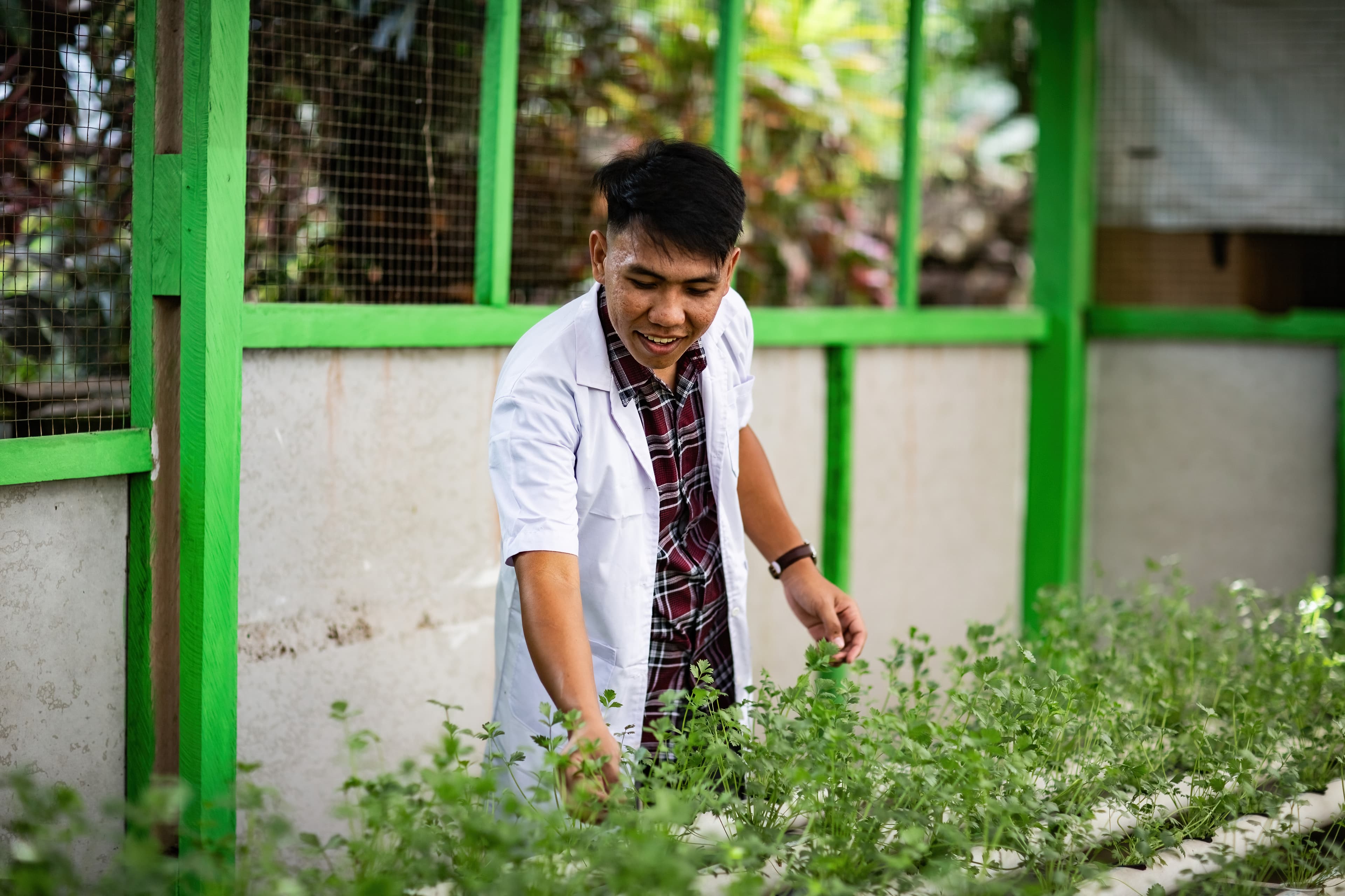 A young man wearing a white lab coat works with plants while smiling.