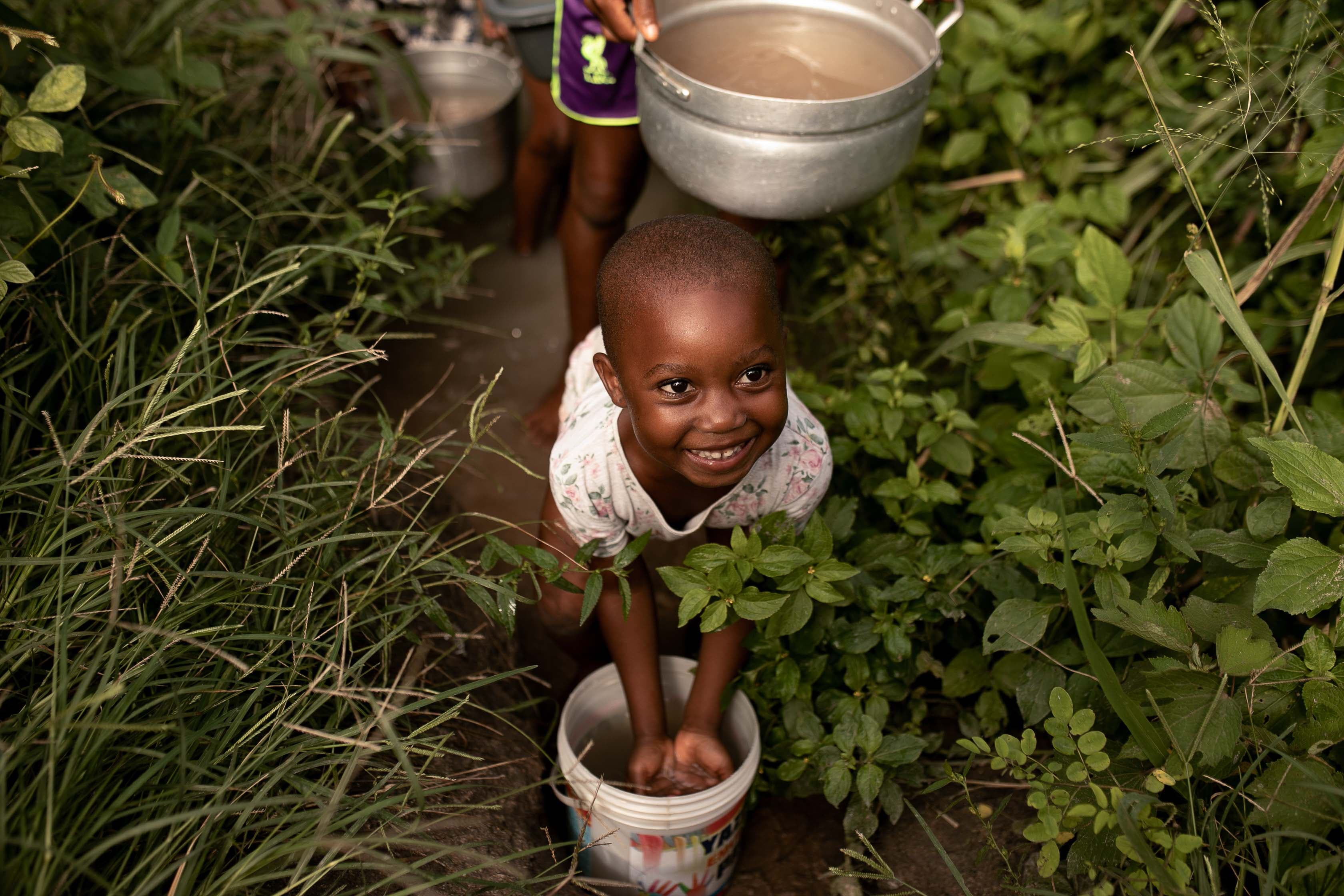 A young African girl places her hands in a bucket of water while smiling for the camera.