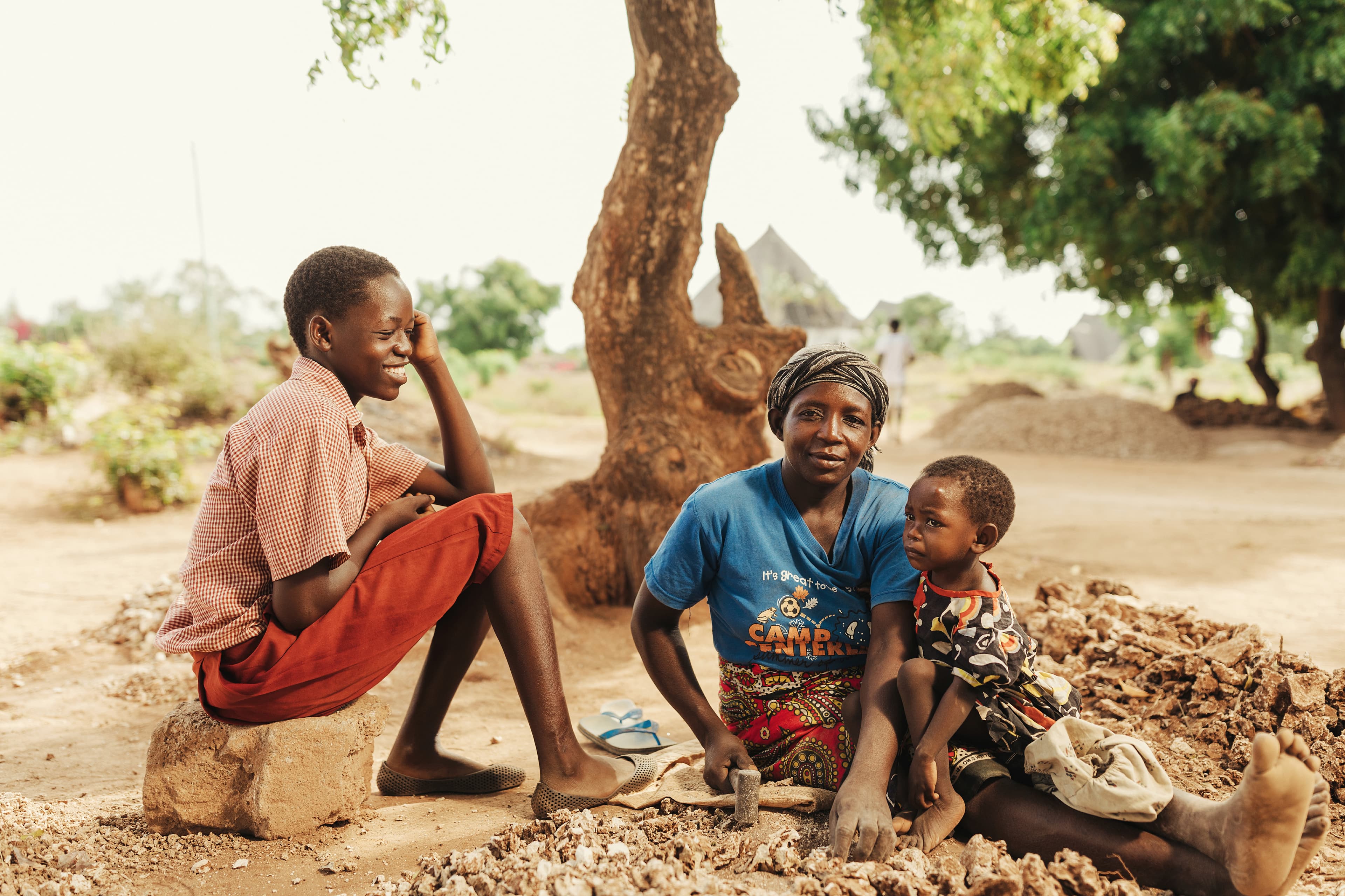 A mother is sitting with her two child as she breaks rocks with a hammer.