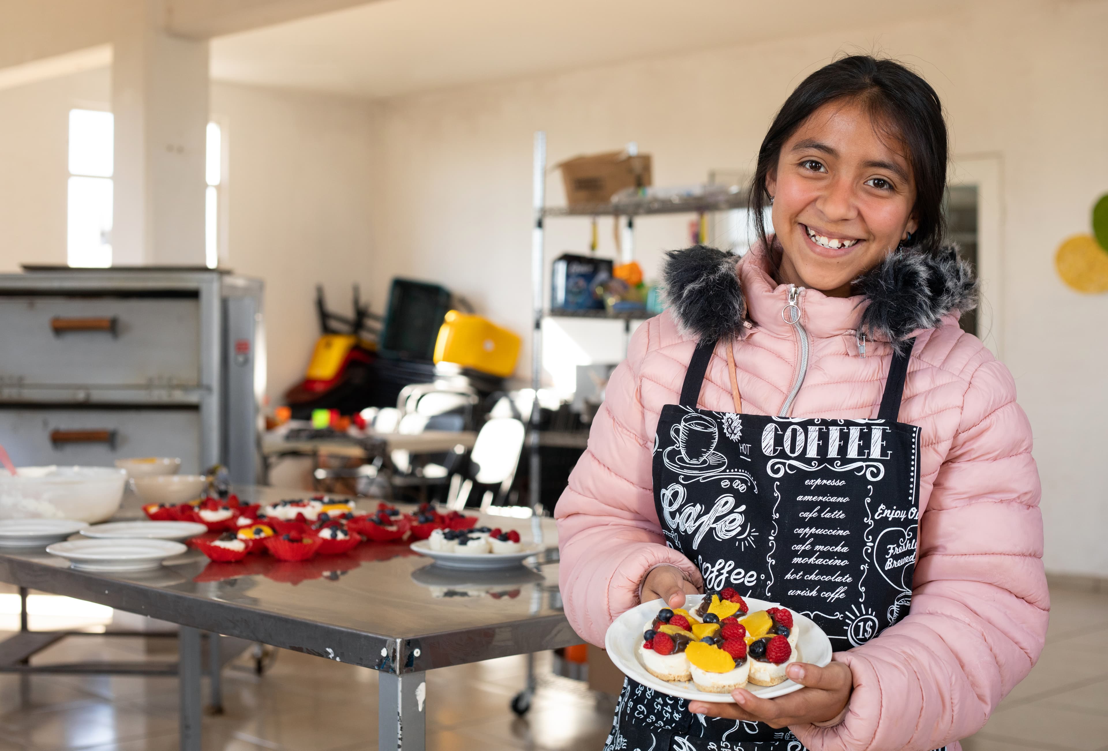 A teenaged girl stands in a kitchen holding cupcakes. She is wearing an apron and pink jacket.