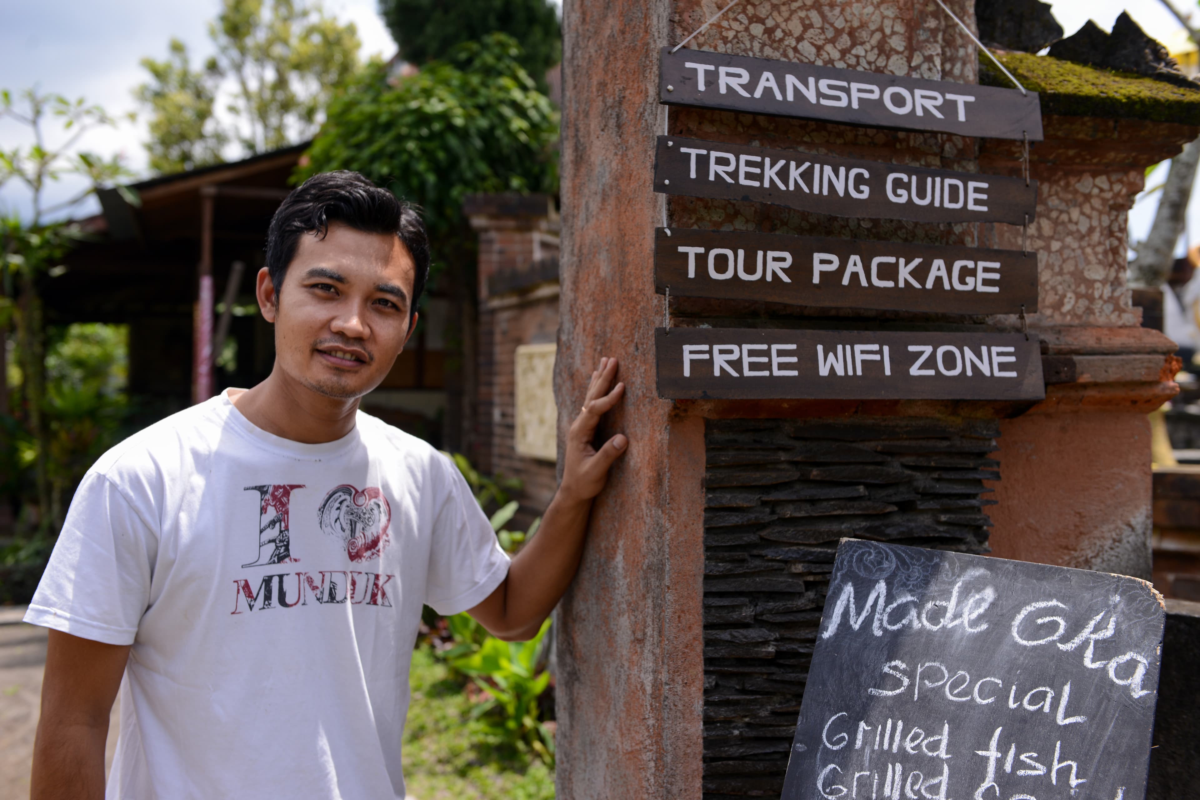 An adult man wearing a white t-shirt stands next to a sign while smiling.