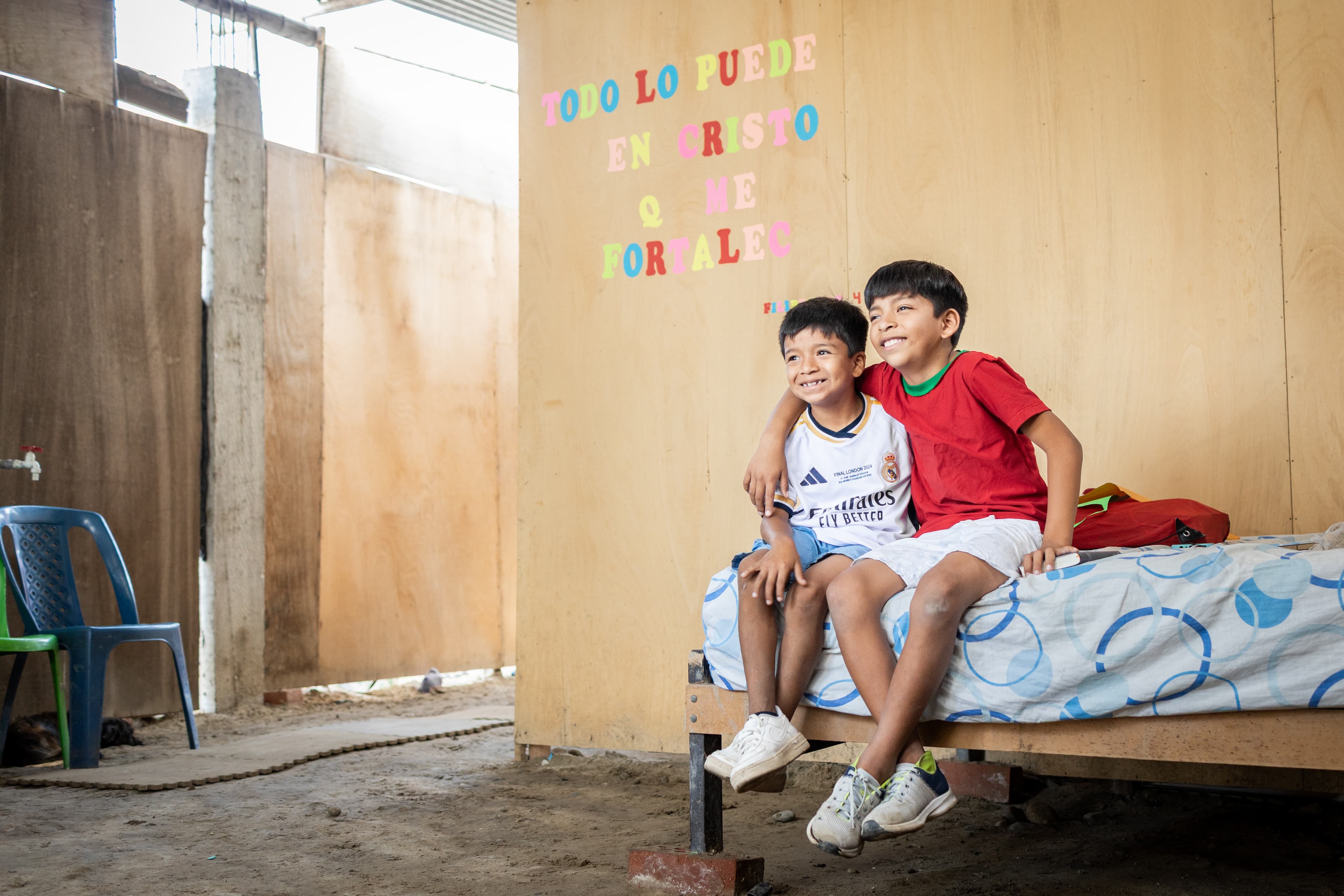 Two boys sit on a bed with a blue and white bedspread in front of a wooden wall decorated with a colorful Bible verse, smiling with their arms around each other.
