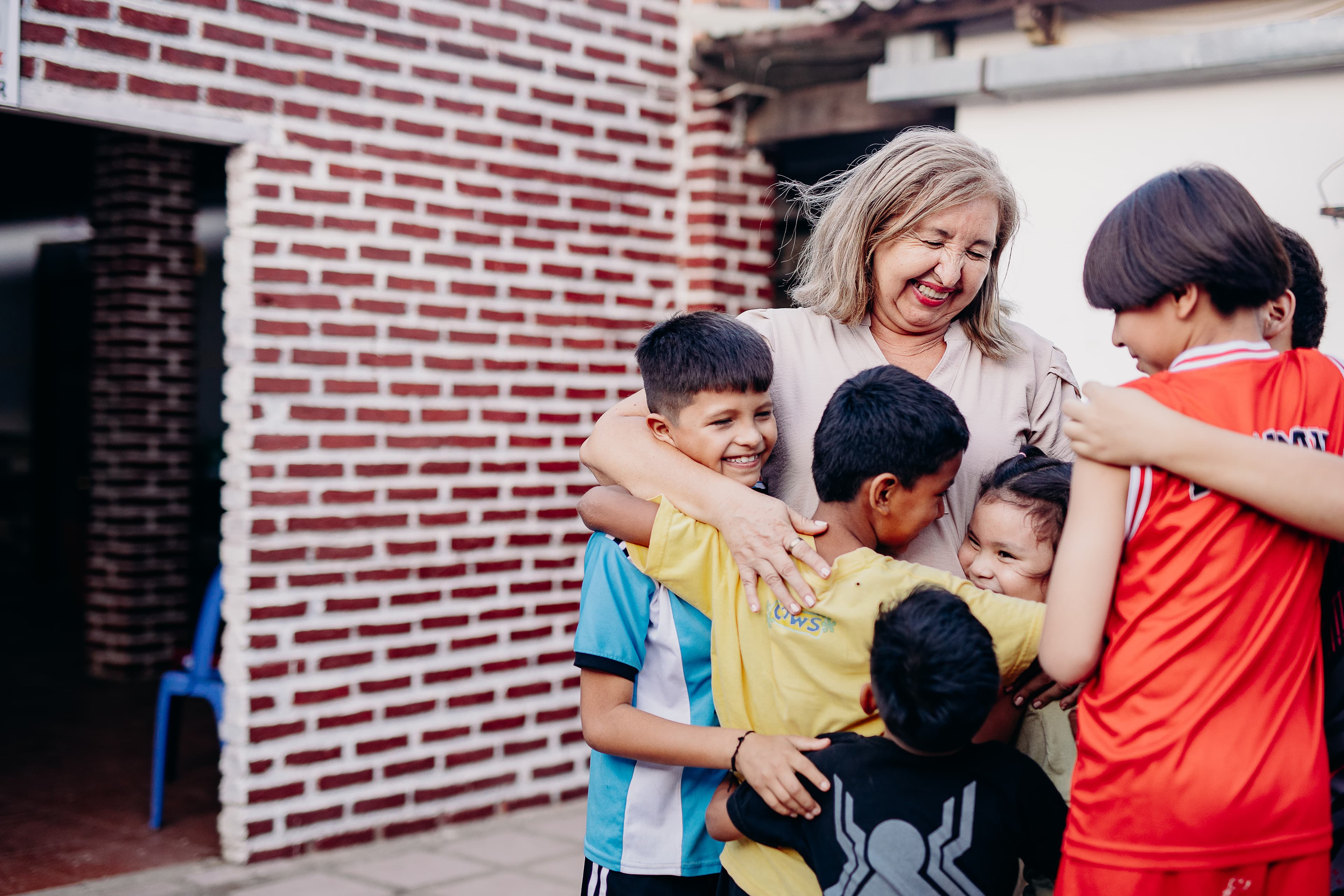 An older woman hugs a group of young boys in front of a brick building.