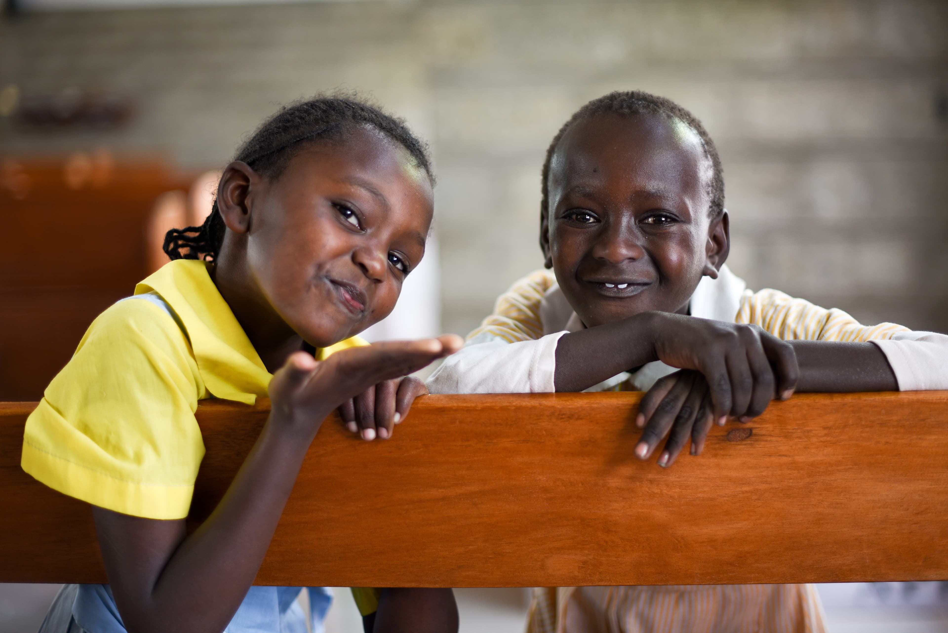 Two African children smile for the camera while sitting on a wooden bench.