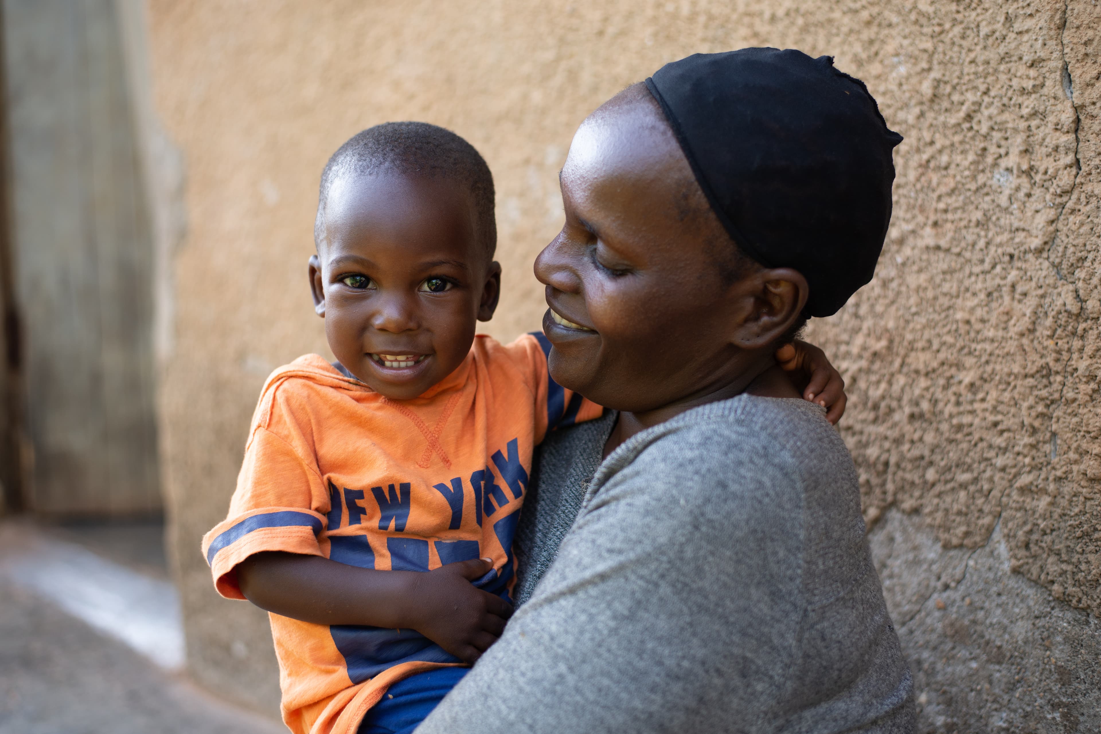 A young African boy smiles for the camera while sitting on his mother's lap.