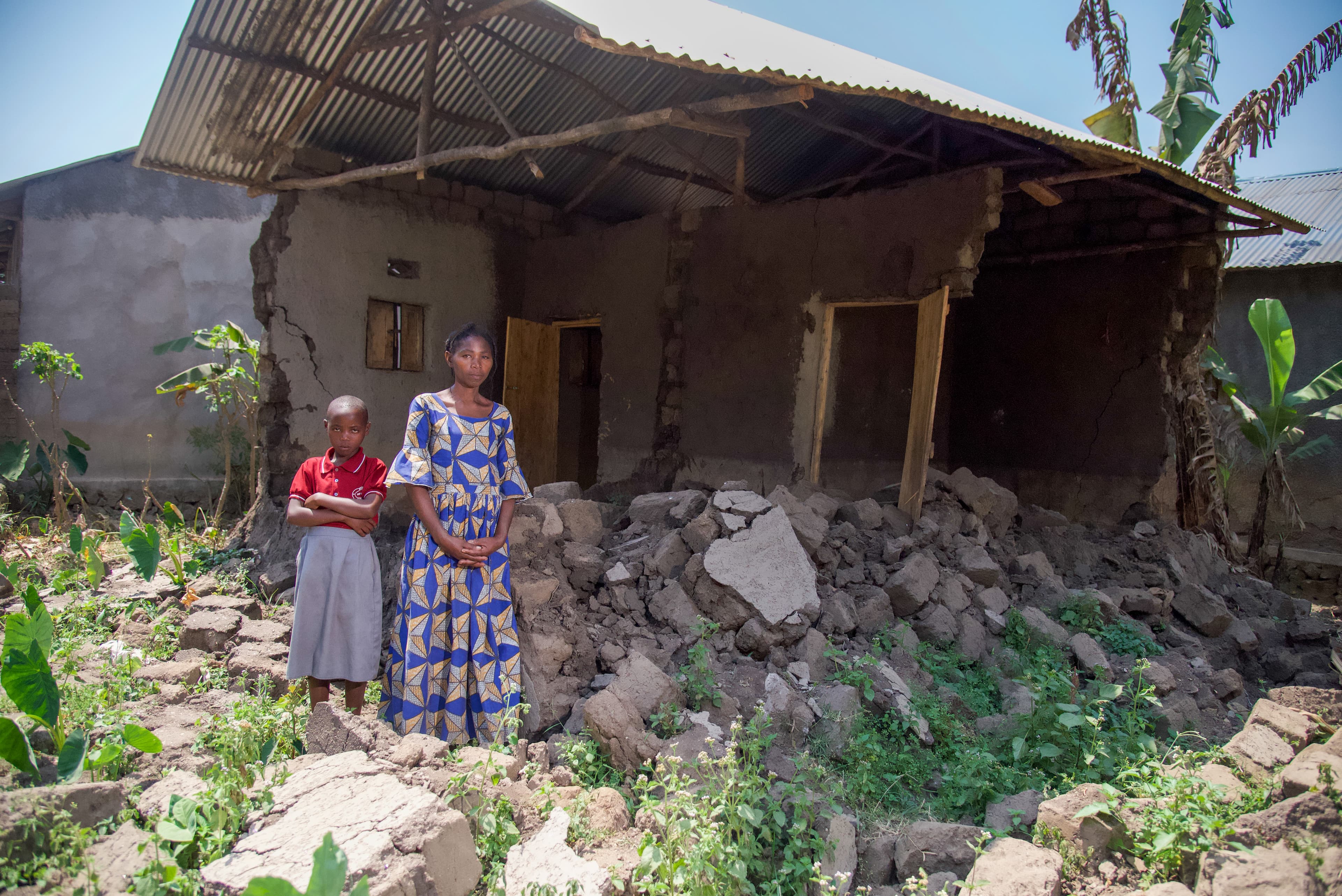 An African woman wearing a patterned dress stands next to a young African girl in front of concrete rubble.