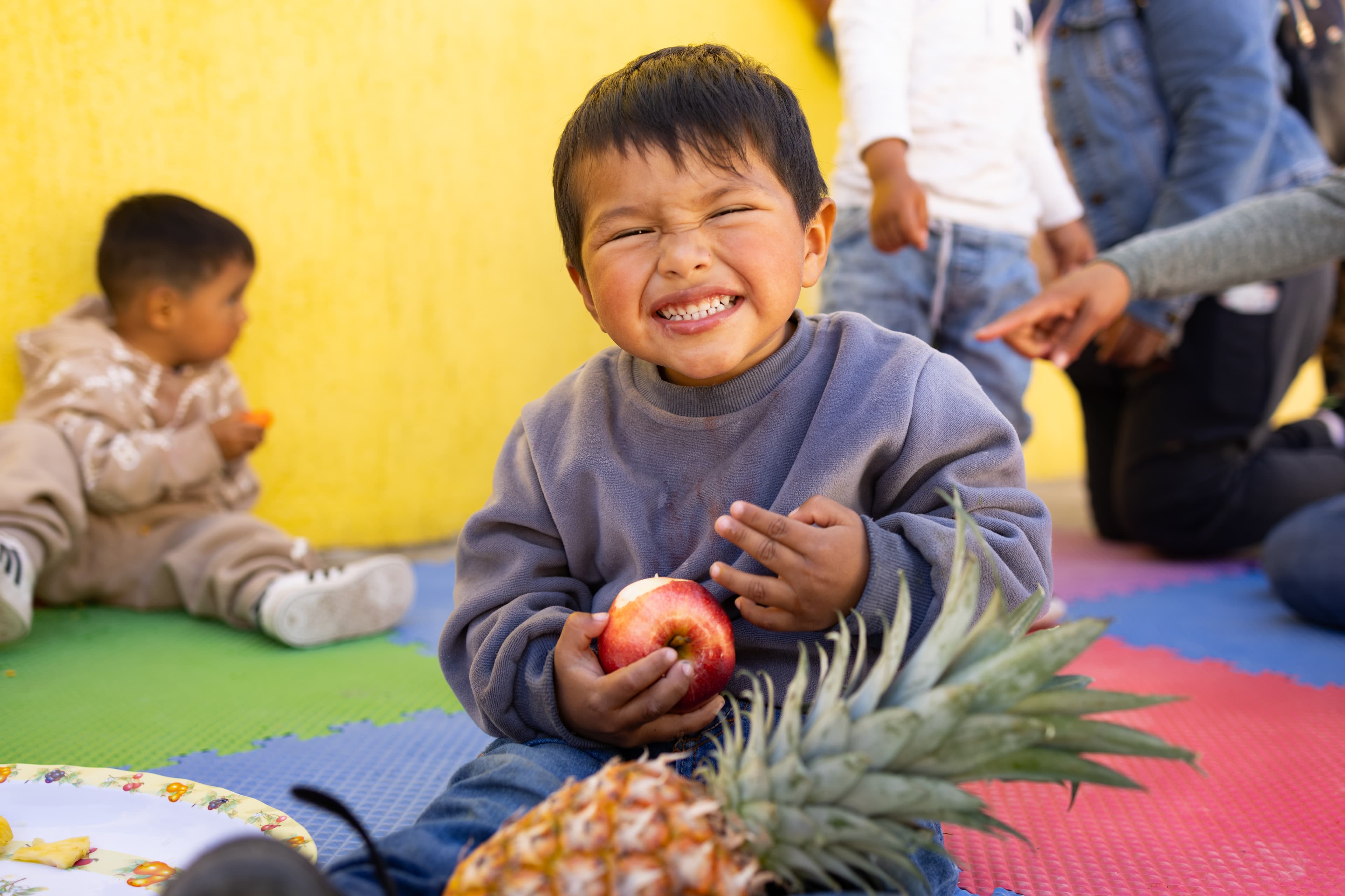 A toddler boy wearing a blue sweatshirt sits and smiles while eating an apple, with a pineapple in his lap.