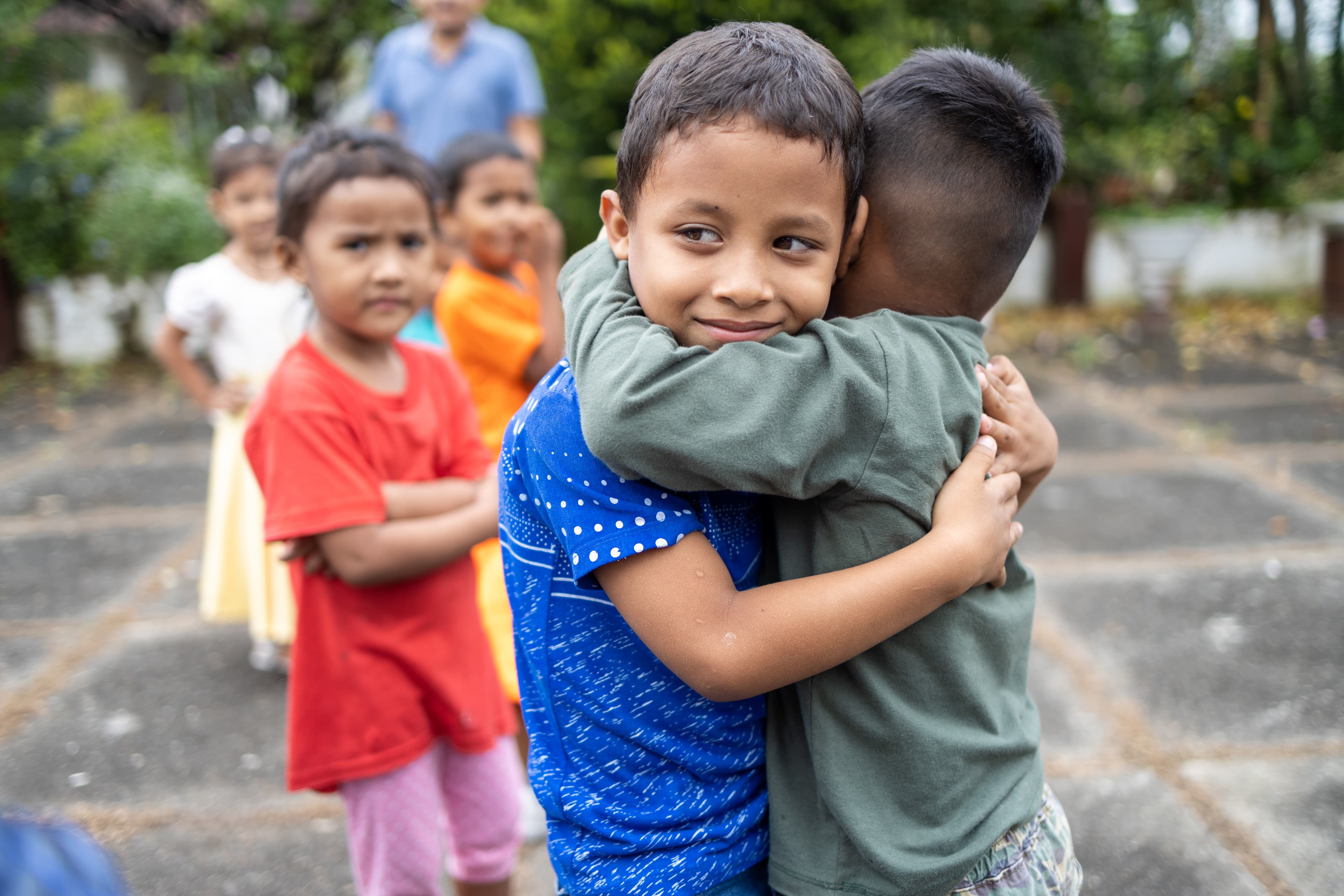 Two young boys, one in a blue shirt and the other in a green shirt, are hugging outside.
