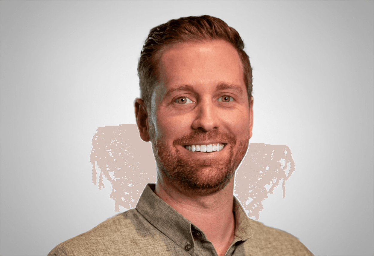 A man in a tan colored button up shirt is smiling, with a grey background behind him.