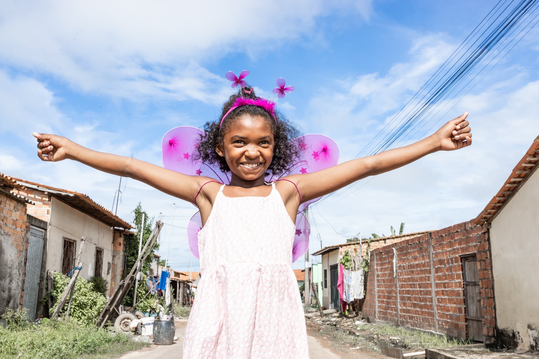 A young girl wearing pink butterfly wings stretches out her arms and smiles.