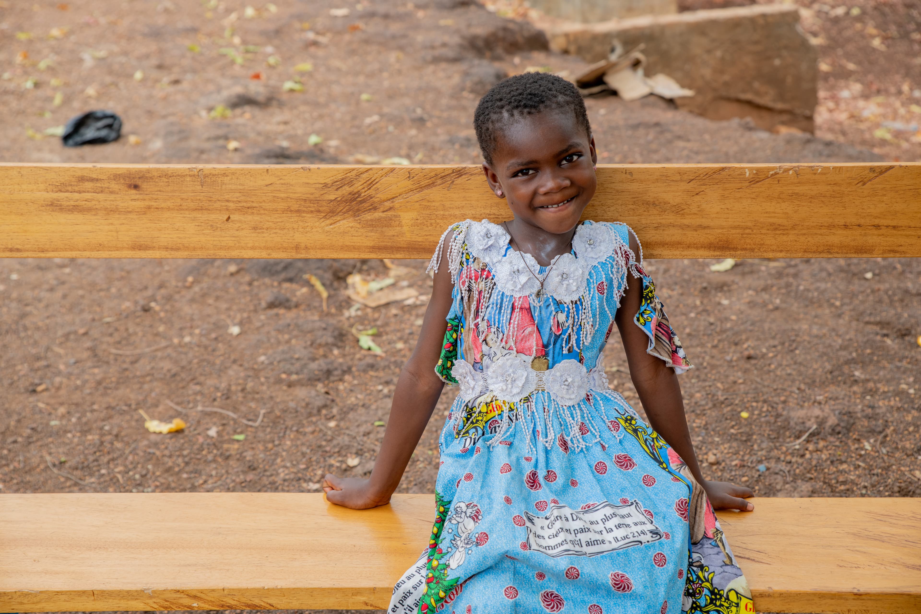 A young African girl wearing a floral dress sits on a wooden bench and smiles for the camera.