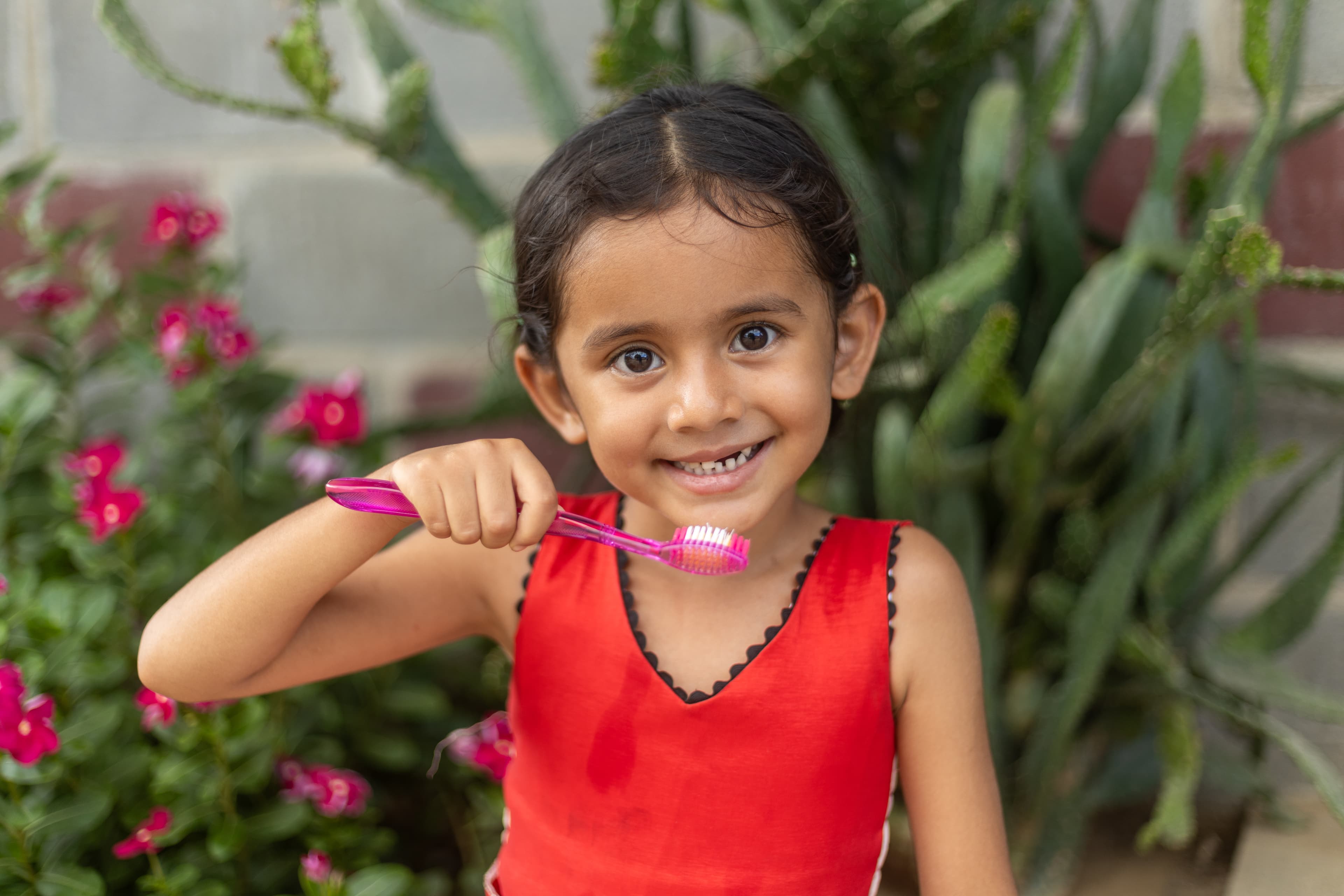 A young girl wearing red smiles at the camera while holding a pink toothbrush.