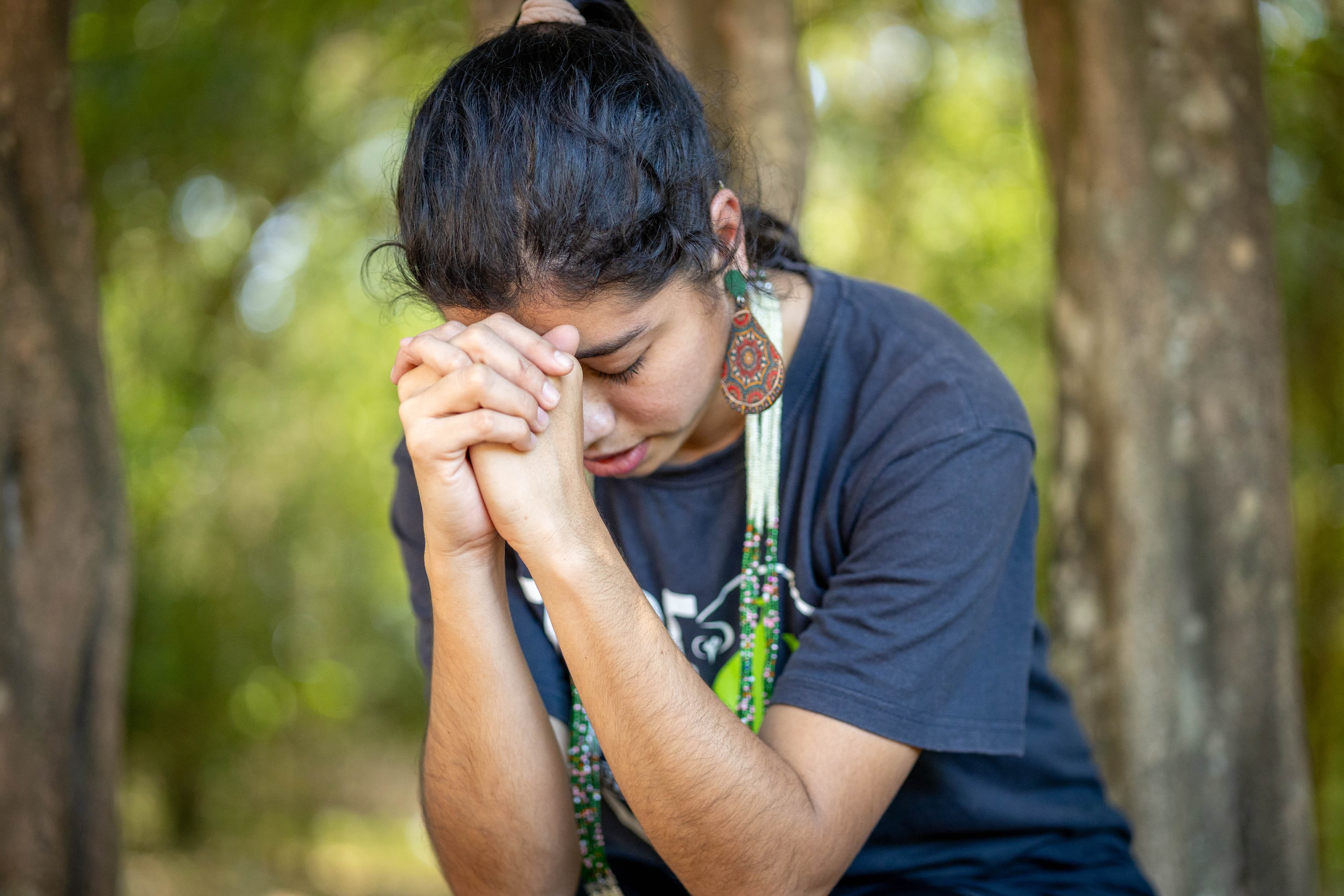 A woman wearing a black shirt and colorful earrings bows her head in prayer. Trees are in the background.