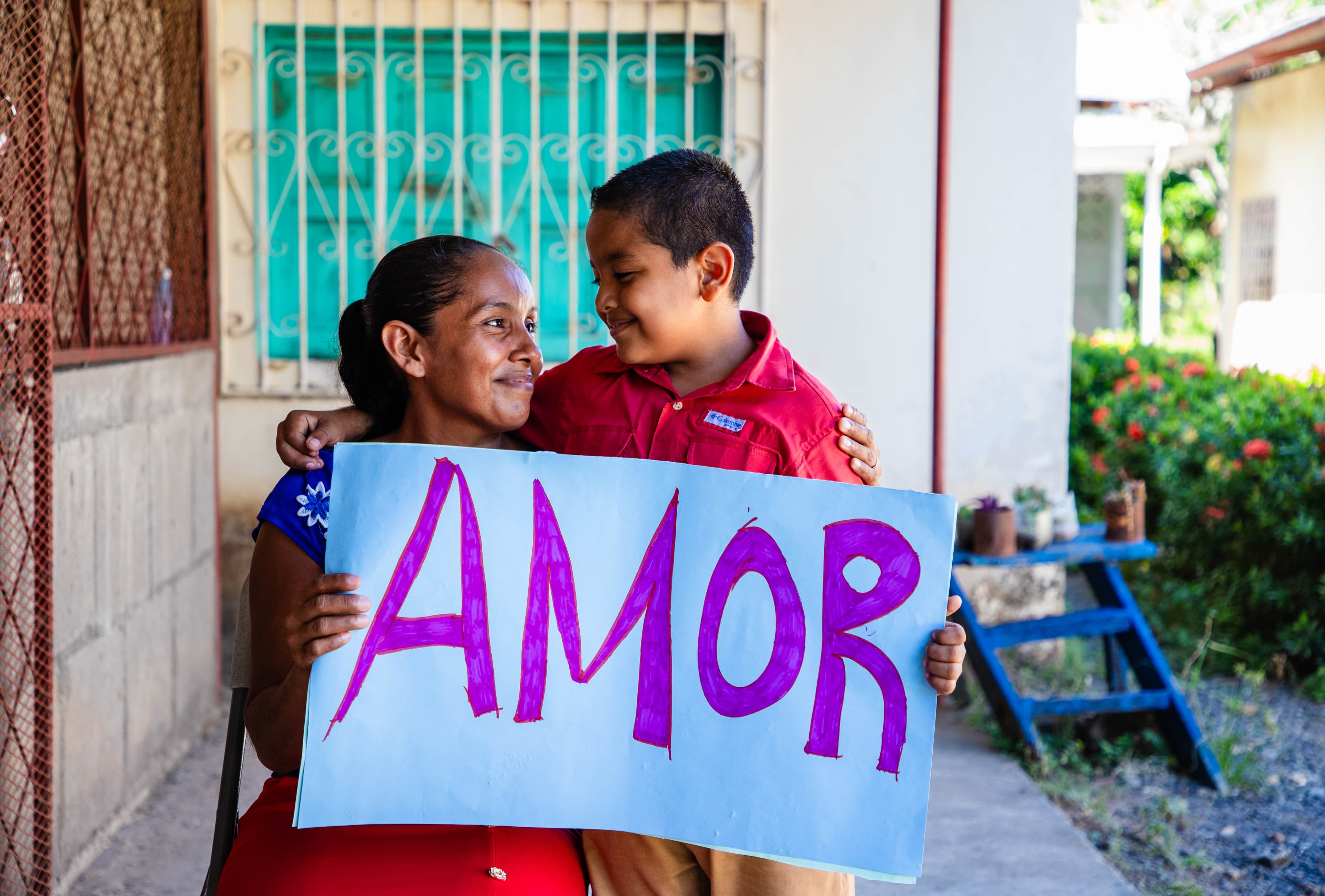 A mom and son hold a sign that says “amor” while smiling at each other.