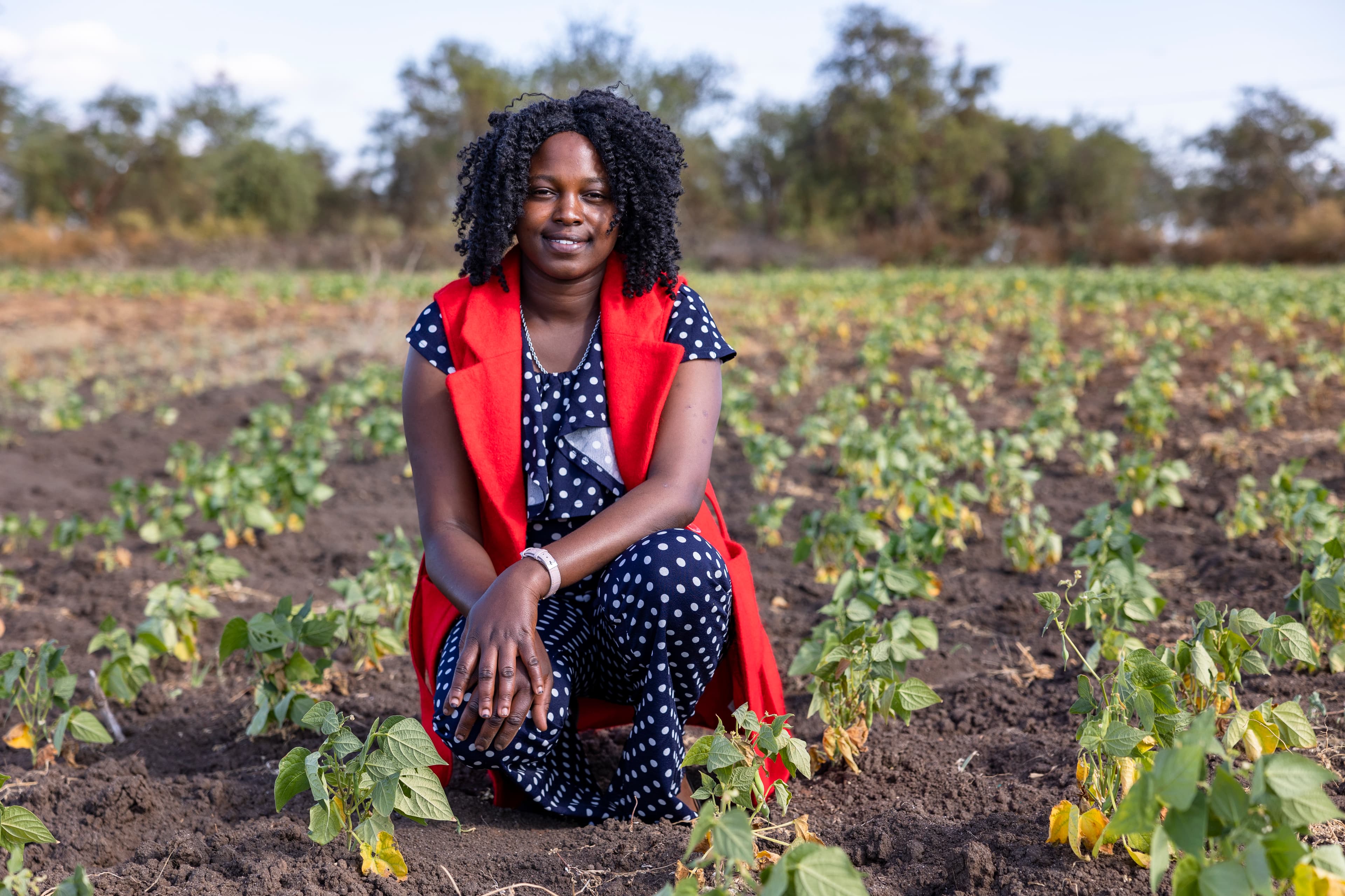 A young African woman wearing a red vest kneels in a field of plants and smiles.