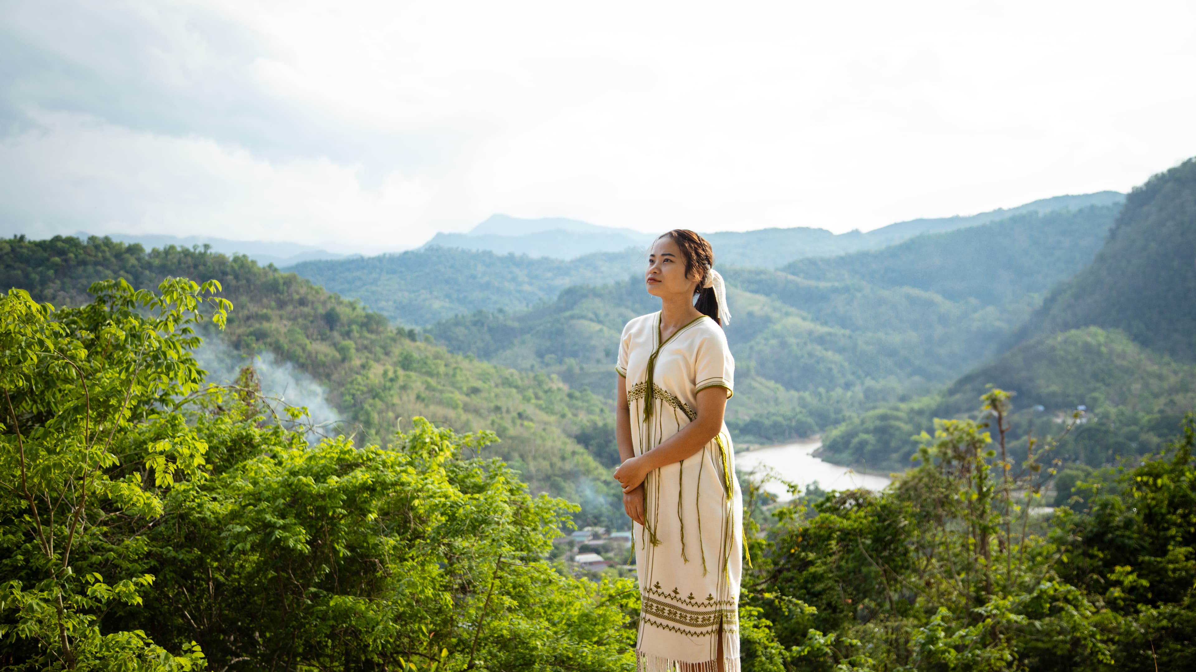 A teenage girl is standing outside on a viewpoint, looking up, with trees, hills and a river in the background.