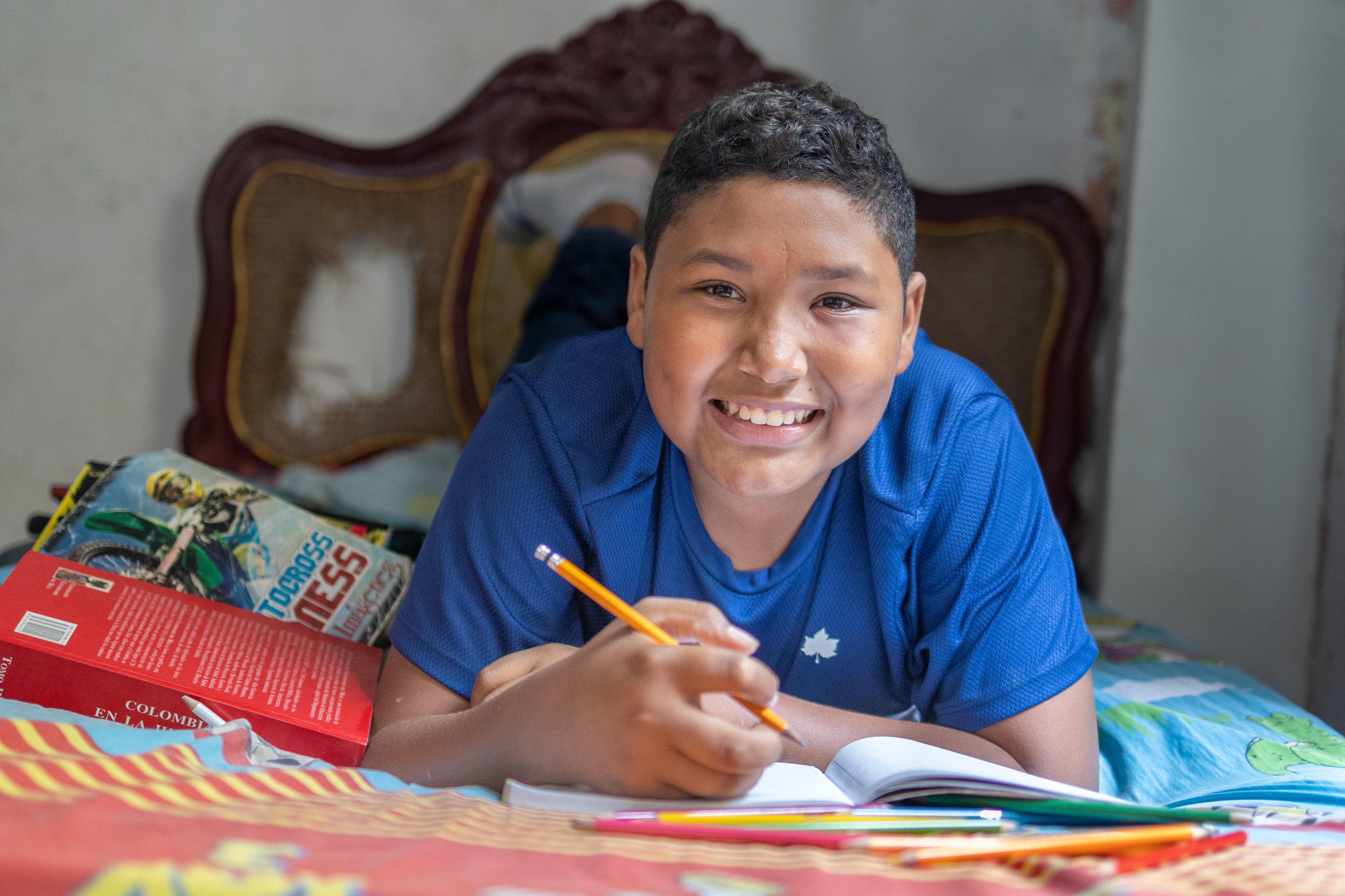 A young boy lays on his bed and writes in a book while smiling.