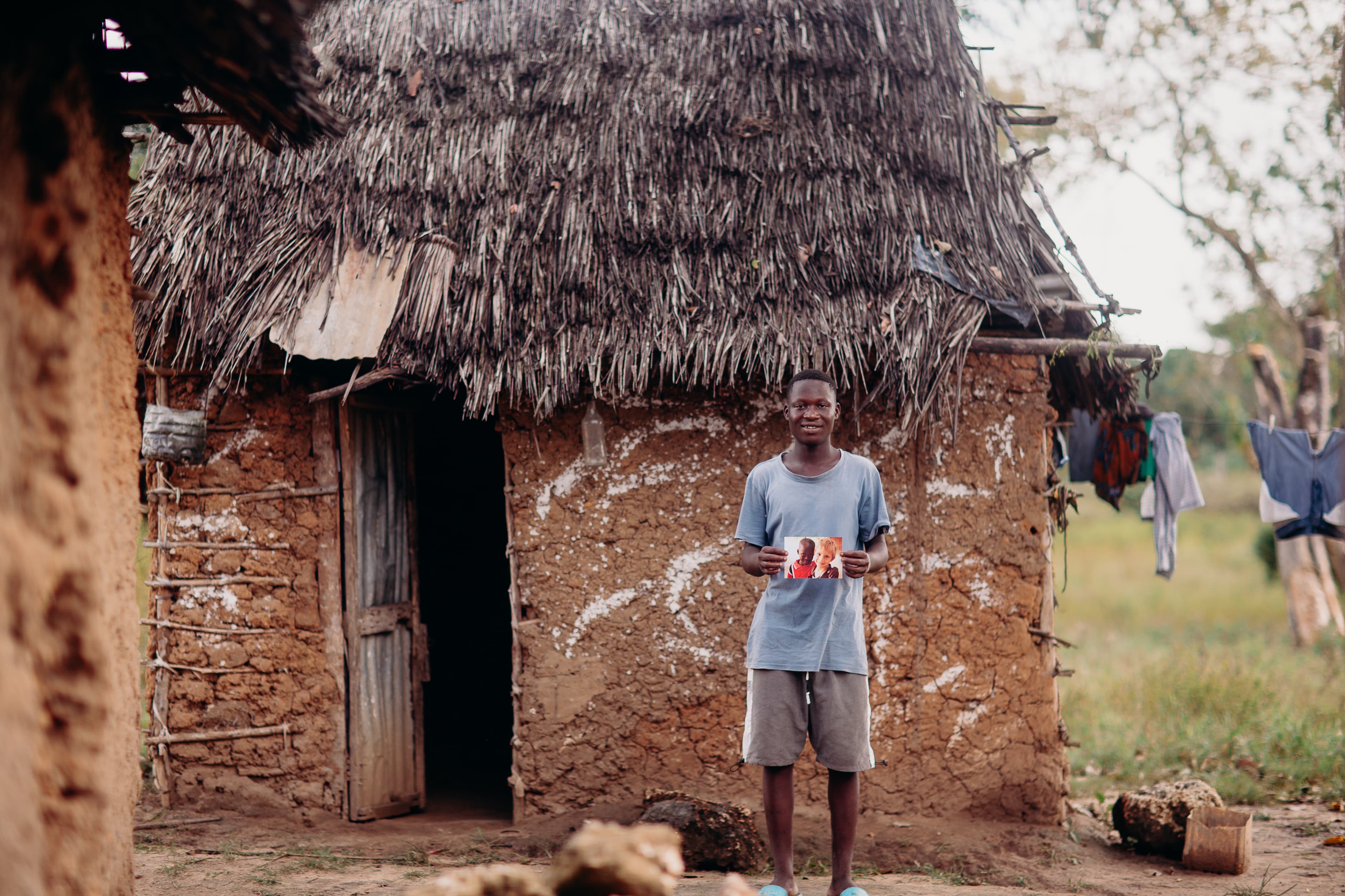 A teen African boy holds a photo while standing in front of a clay home.