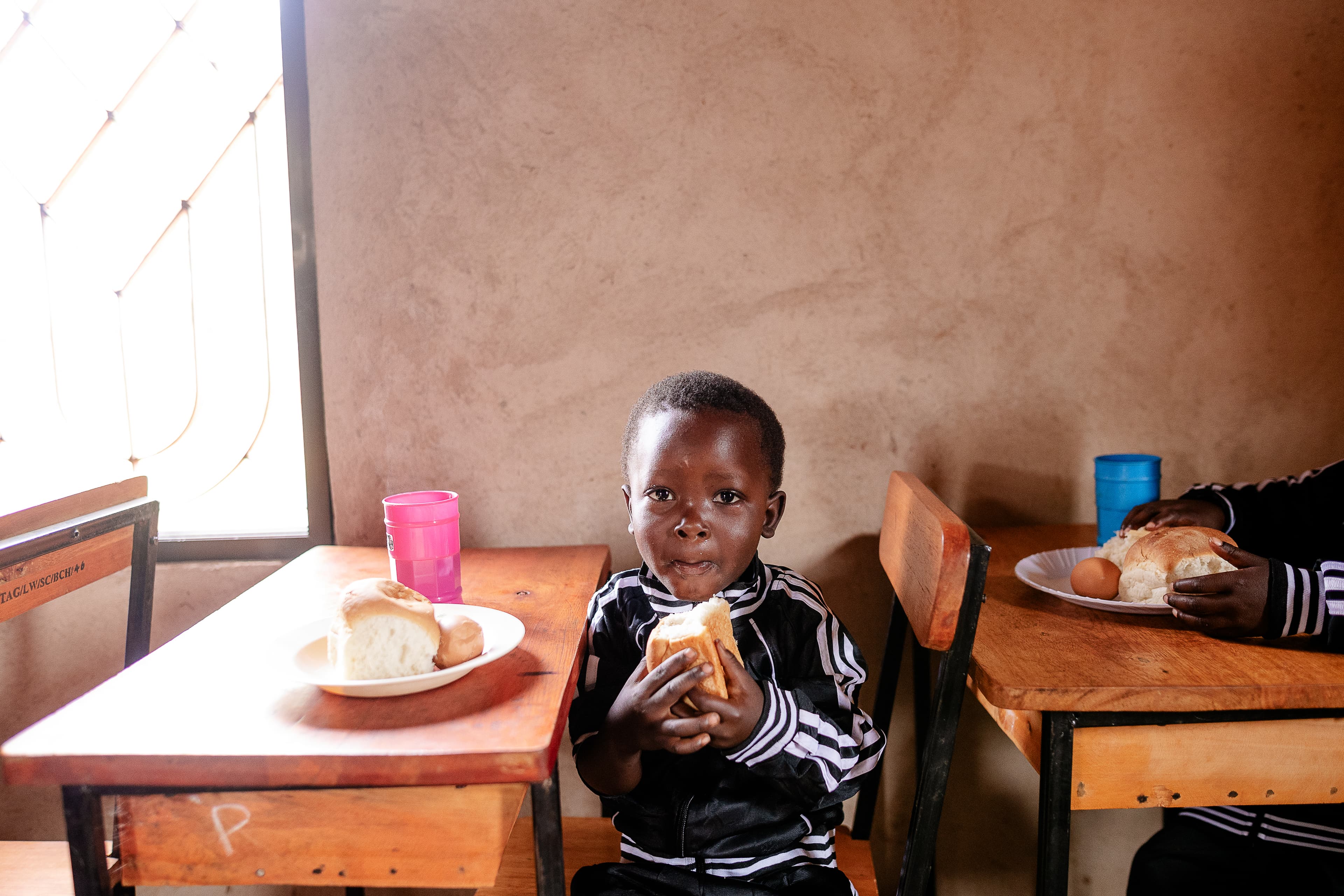 A young child is sitting at a desk eating bread and looking at the camera.