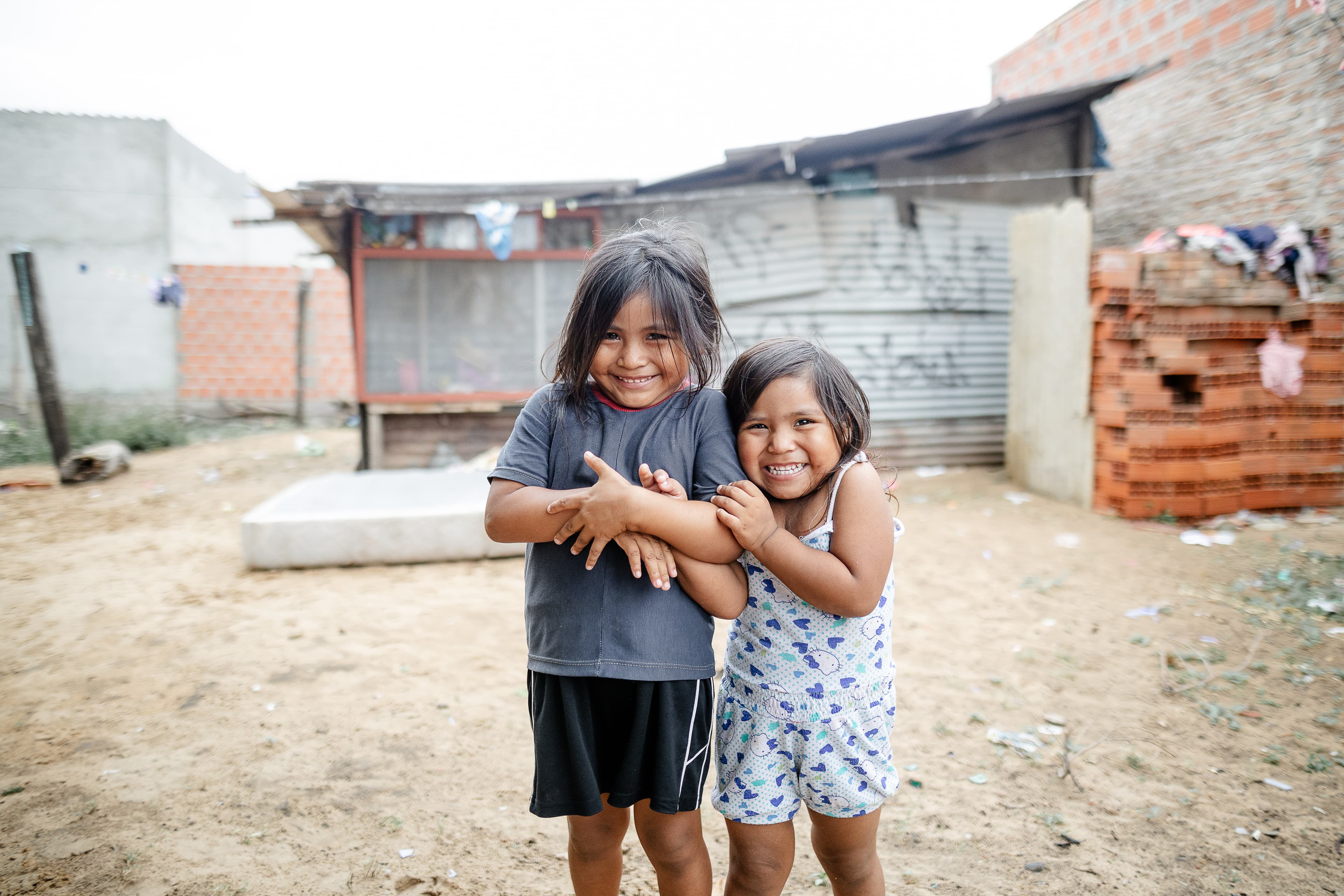 Two playful girls embrace each, one in a gray shirt and skirt, the other in a white floral top and shorts. A makeshift shelter is behind them.