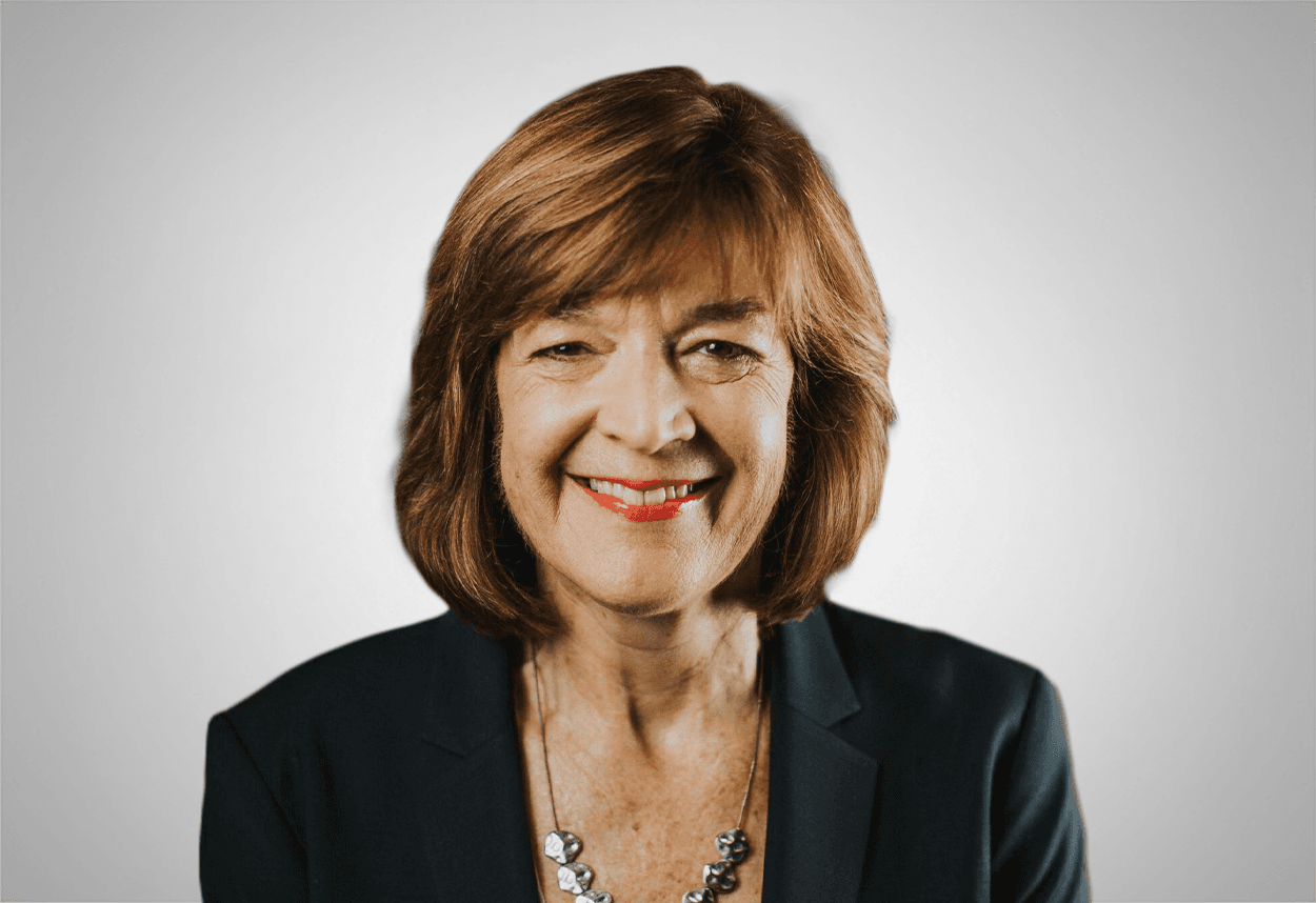 A woman in a black blazer and silver necklace is smiling, with a grey background behind her.