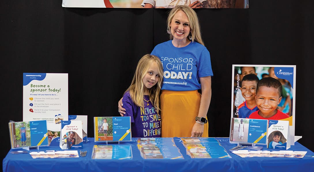 A woman and young girl stand behind a table with signs saying, "Become a sponsor today!"