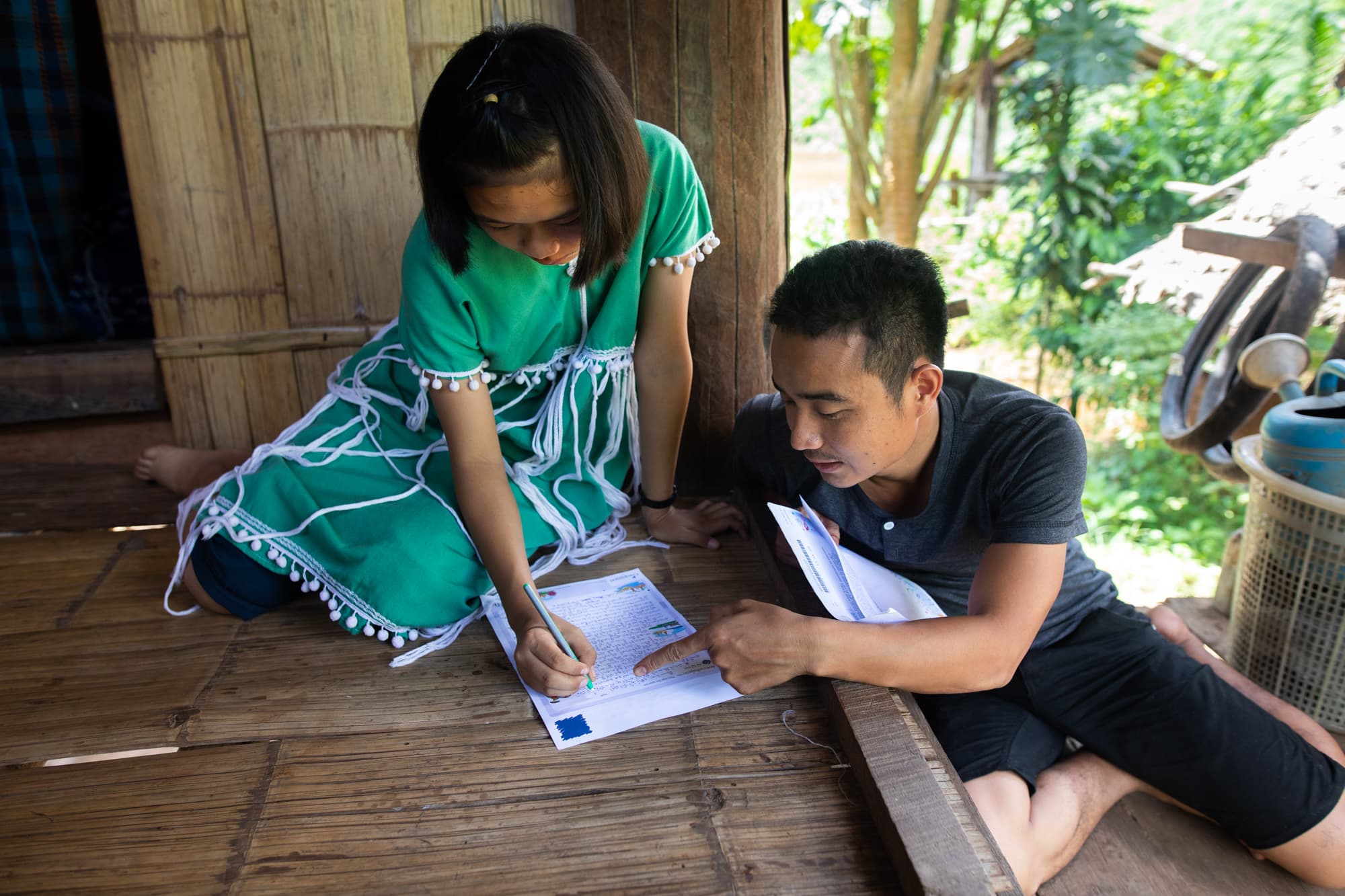 A Project Director is sitting and helping a young girl write a letter to her sponsor.