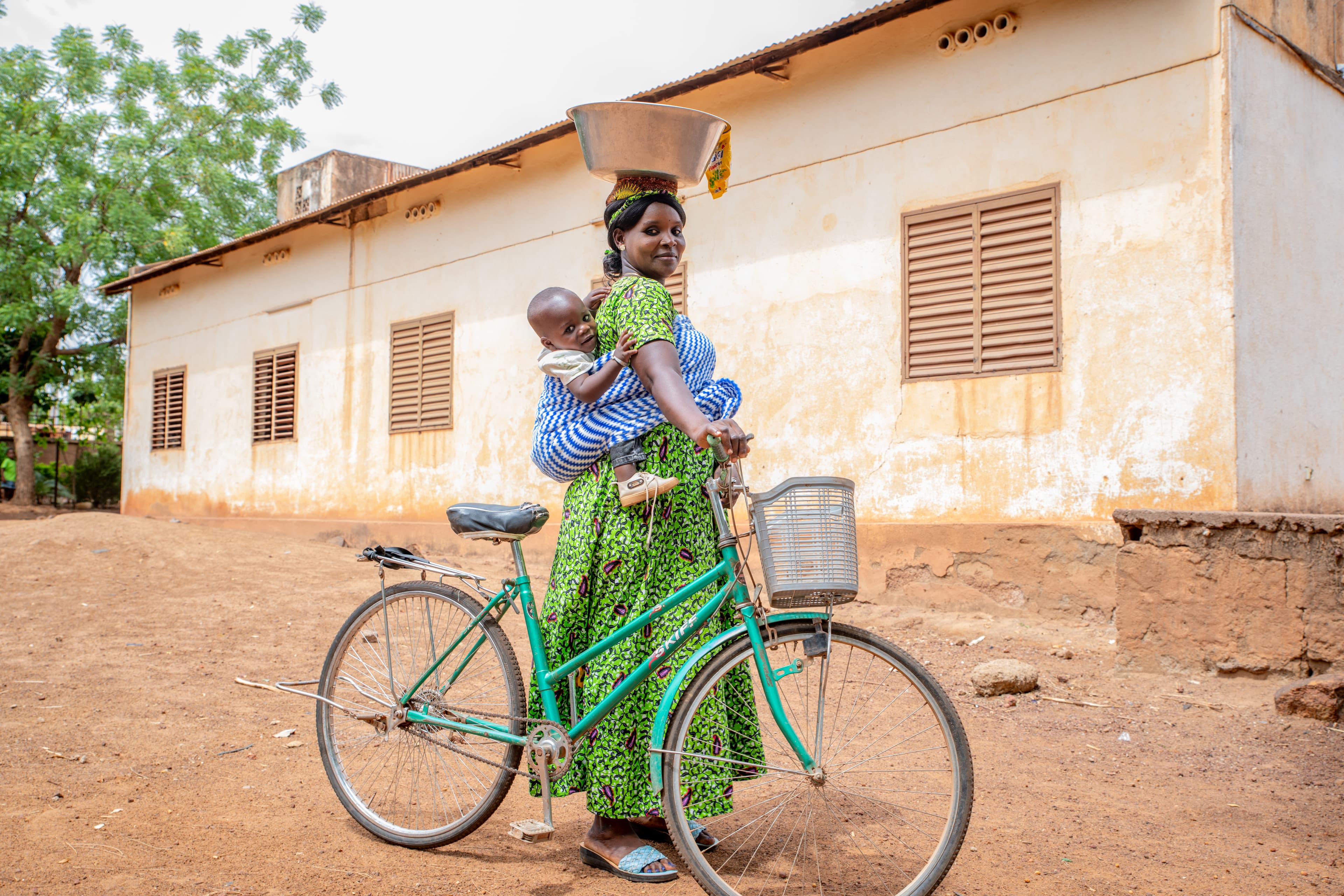 A mother is standing with a bicycle, with her baby wrapped around her back and a plate on her head.