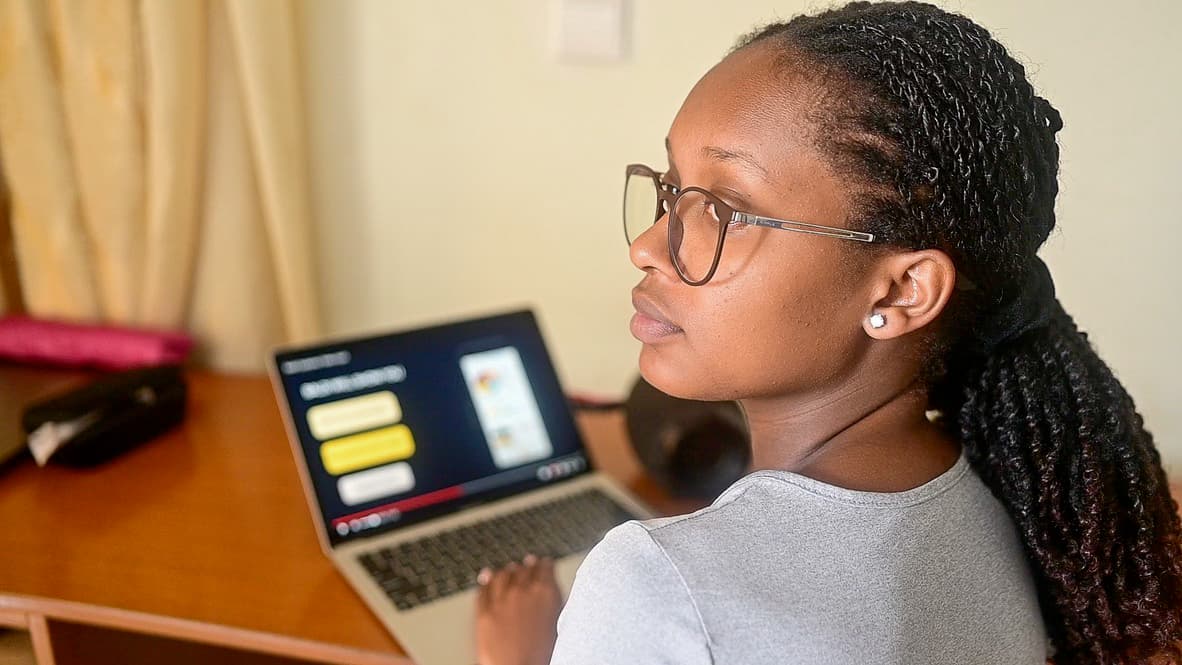 A teen girl sits at an open laptop at a desk. Her hair is twisted and she is wearing glasses.