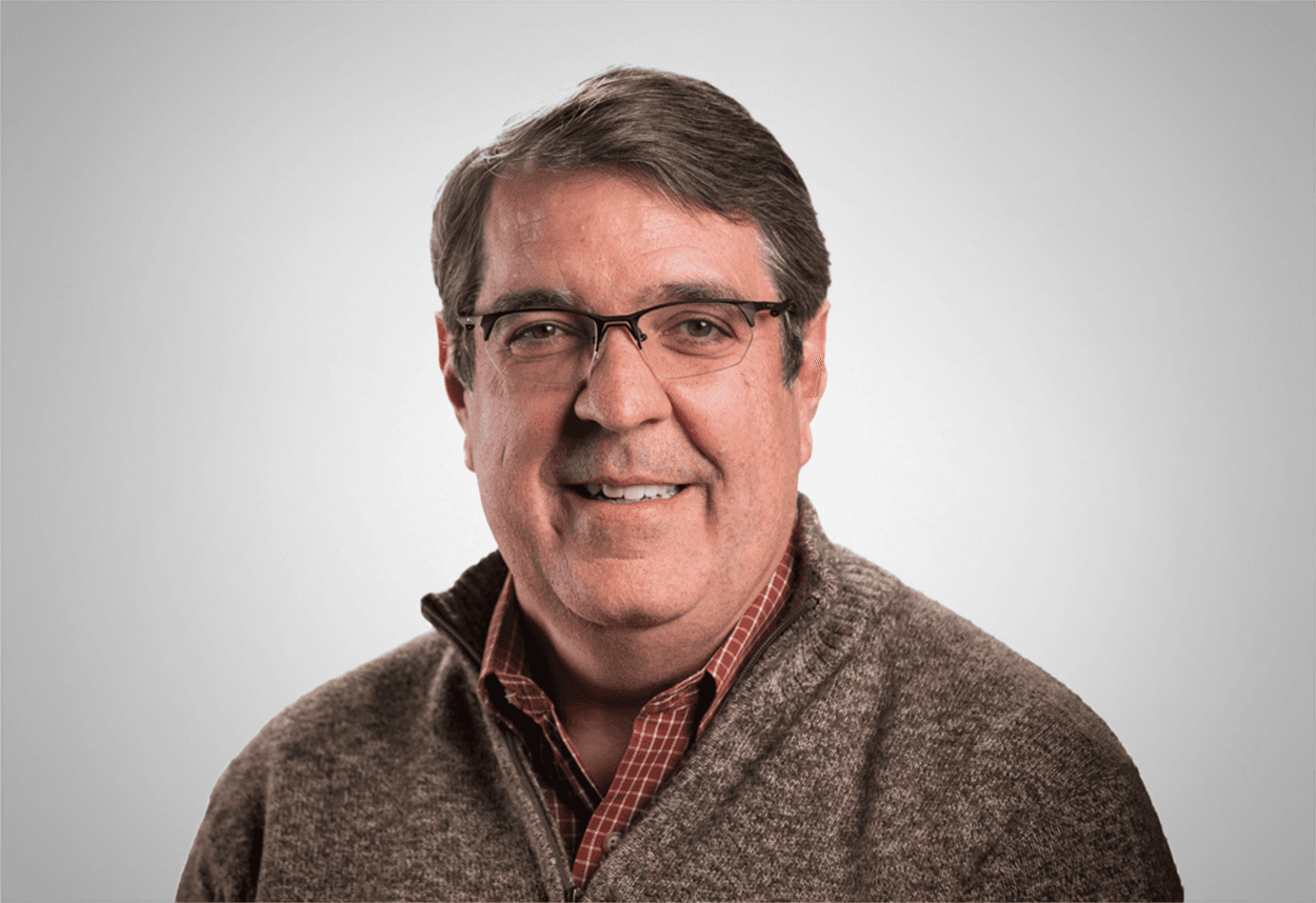 A man in a red checkered shirt, brown sweater and glasses is smiling, with a grey background behind him.