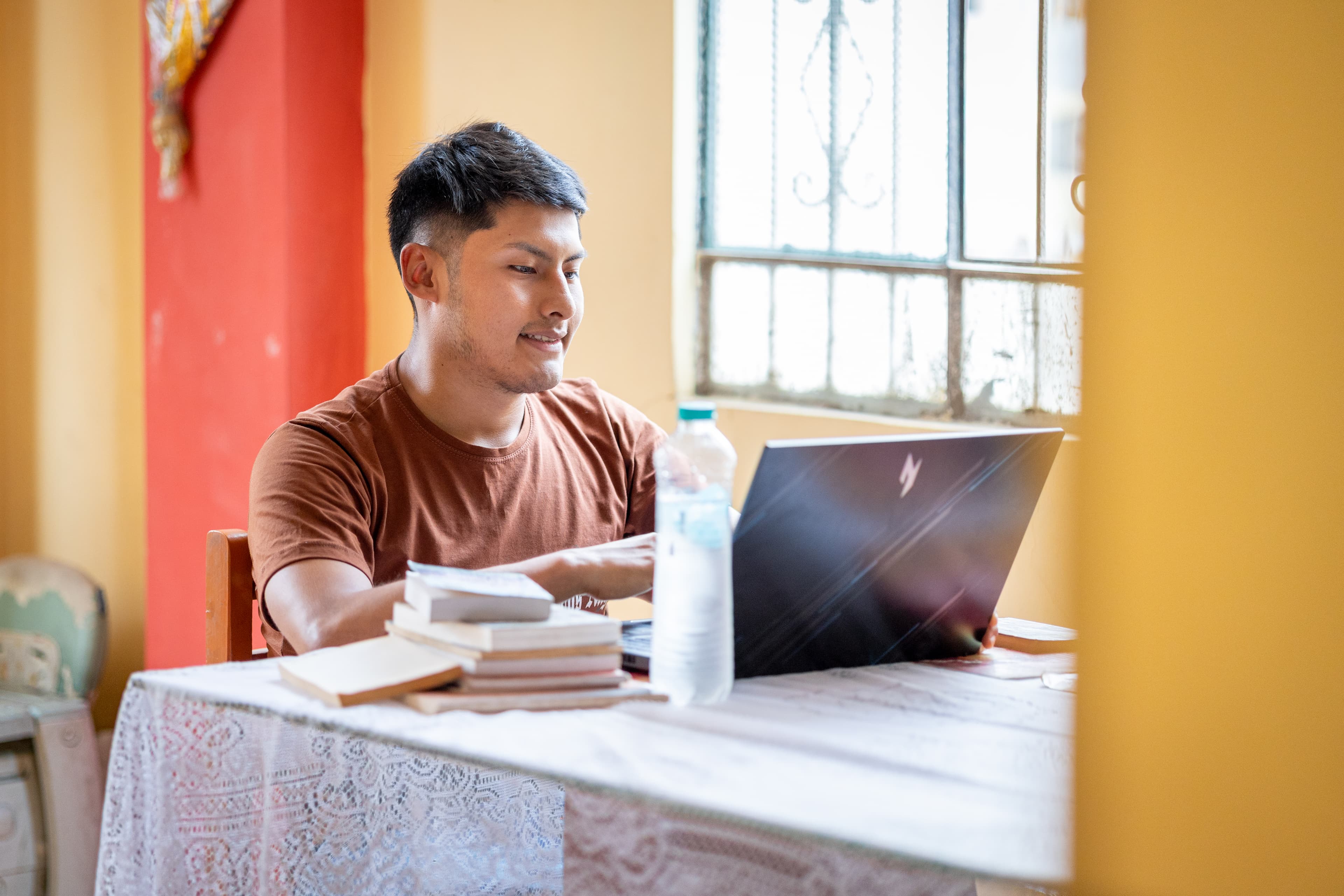 A young man in a brown shirt sits at a white tablecloth-covered table, using a black laptop to connect to a virtual class.