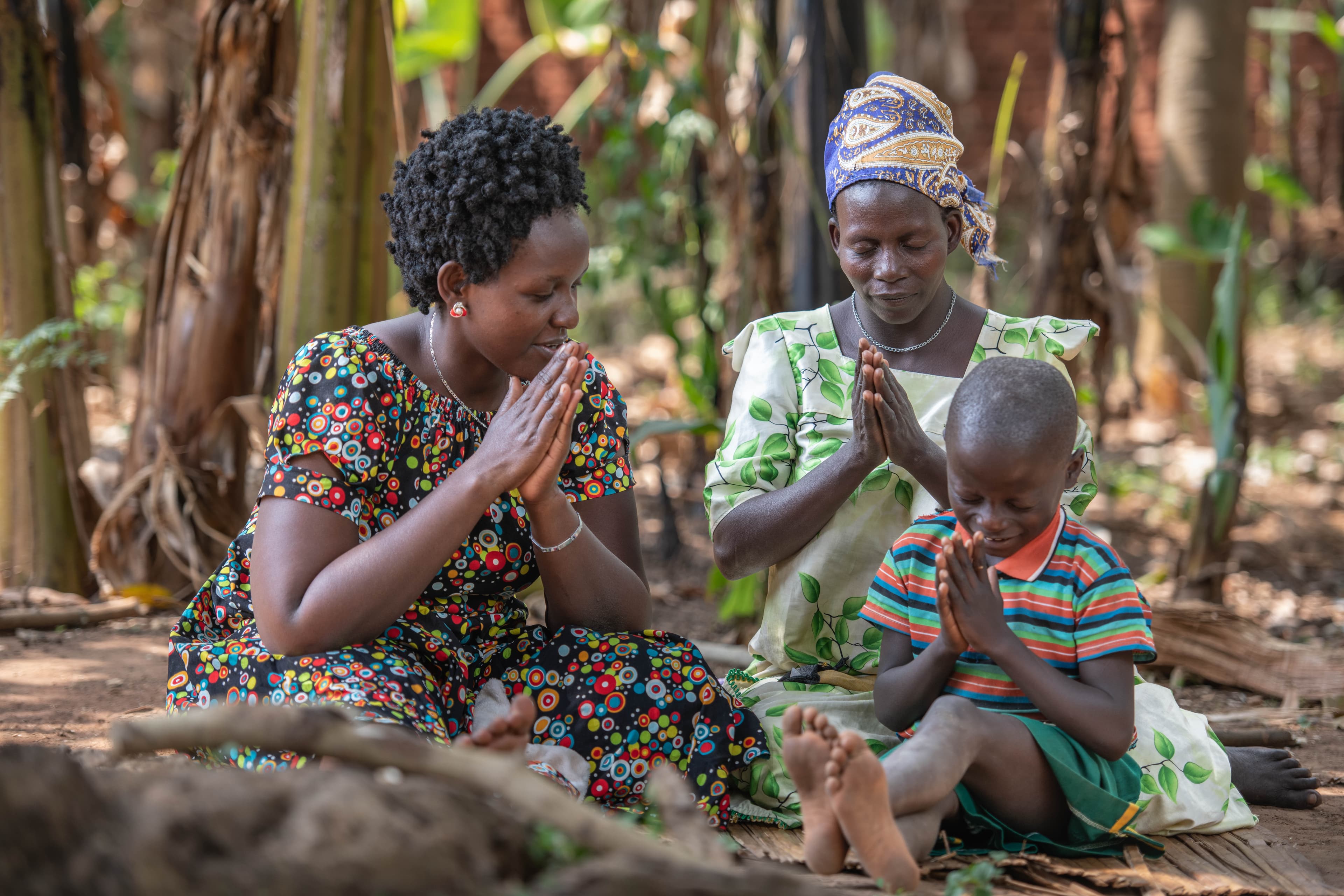 Two African women wearing brightly patterned dresses pray with a young boy.